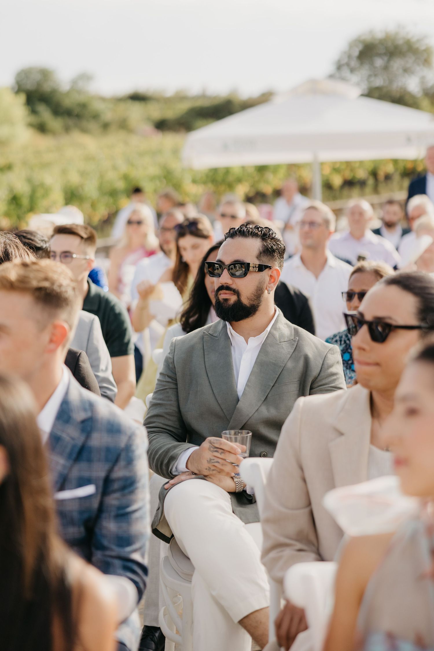 Wedding guests in formal attire seated under white tents at an outdoor ceremony.