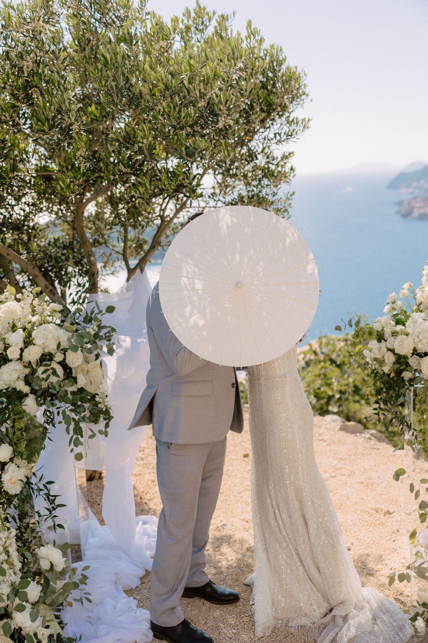 A wedding couple embraces under an olive tree with white floral arrangements.
