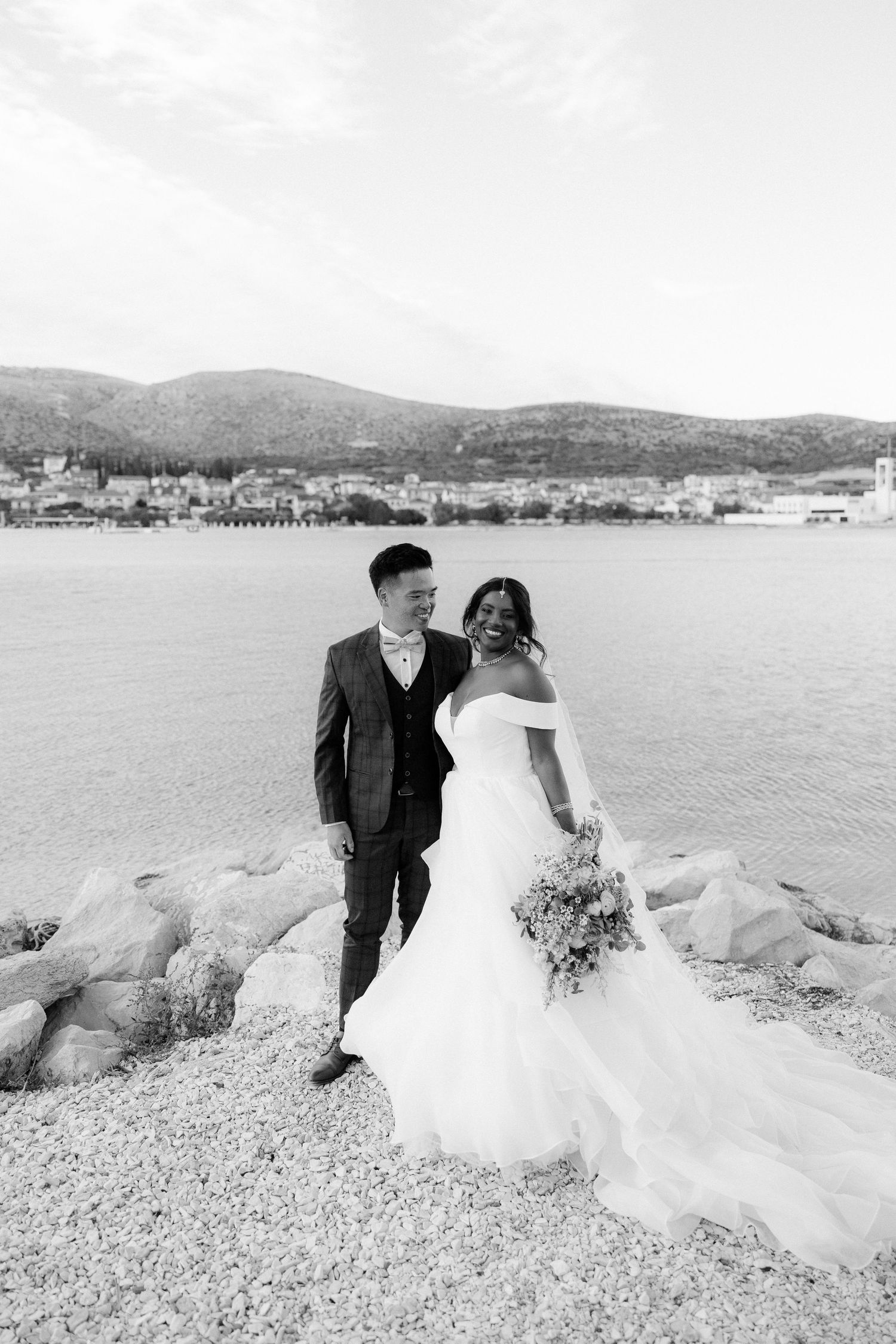 Black and white portrait of newlywed couple standing together on rocky shore with lake and mountains behind.