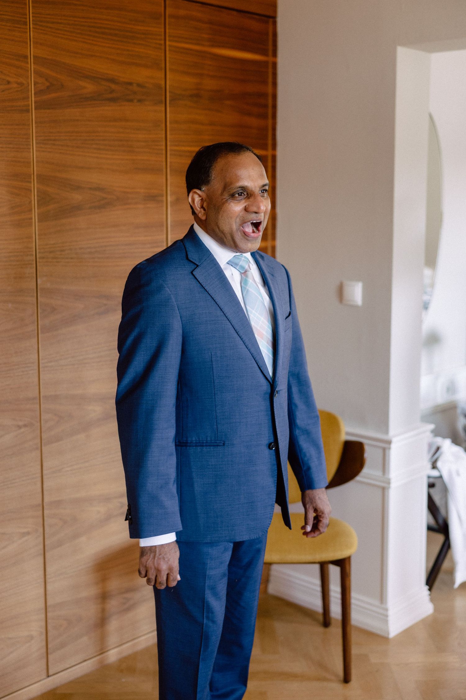 Groom in a blue suit with light blue tie laughing joyfully while getting ready for his wedding day.
