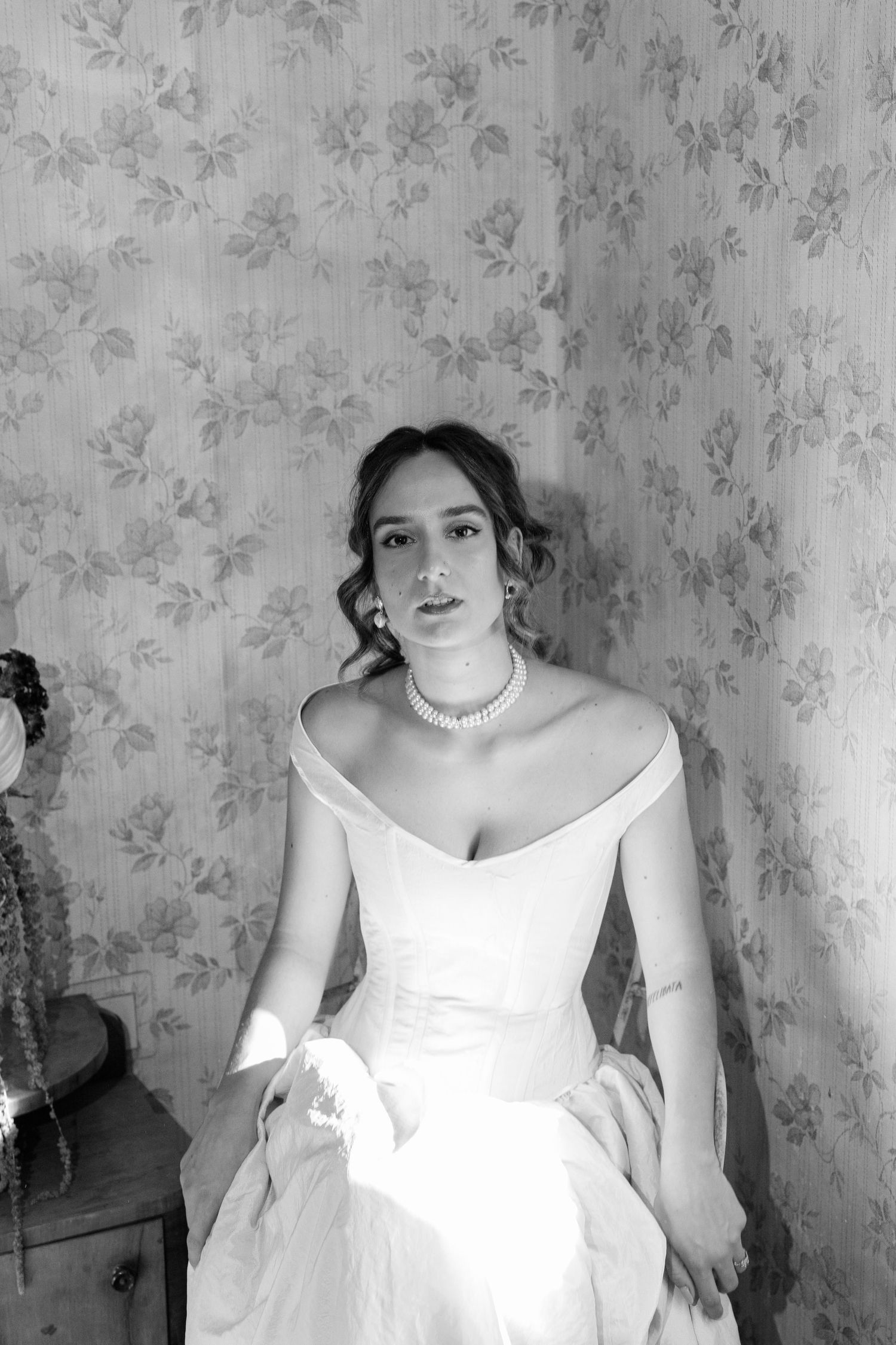 Elegant white wedding dress against floral wallpaper in classic bridal portrait style.