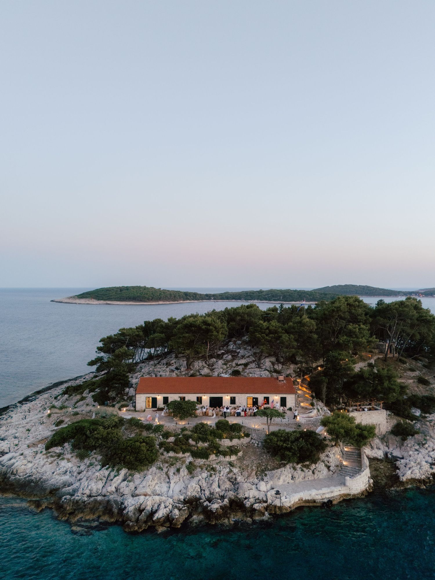 Aerial view of a rustic wedding venue on a rocky Mediterranean island surrounded by turquoise waters at dusk.