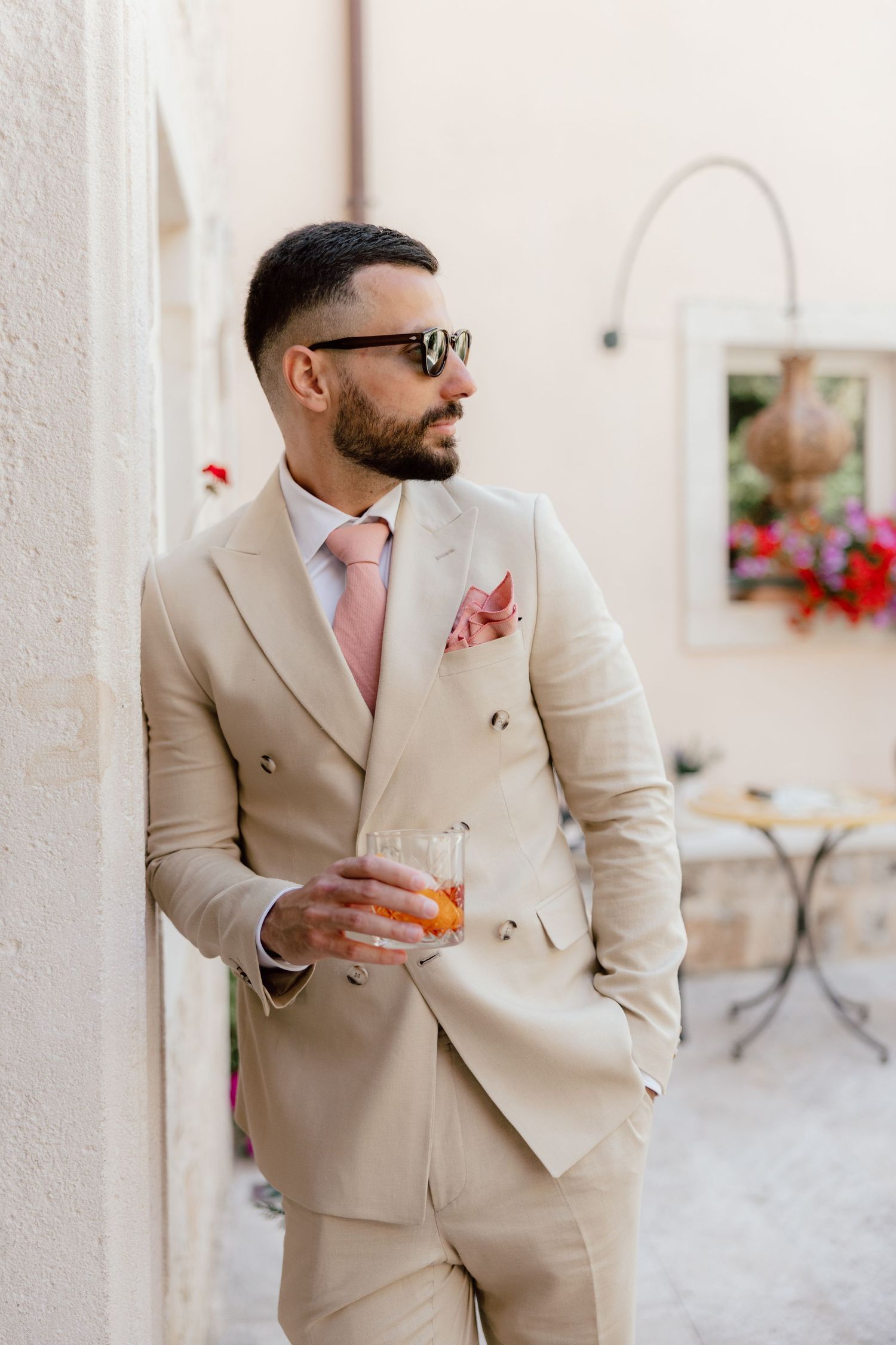 Elegant man in beige double-breasted suit, sunglasses, pink tie, holding a drink outdoors.