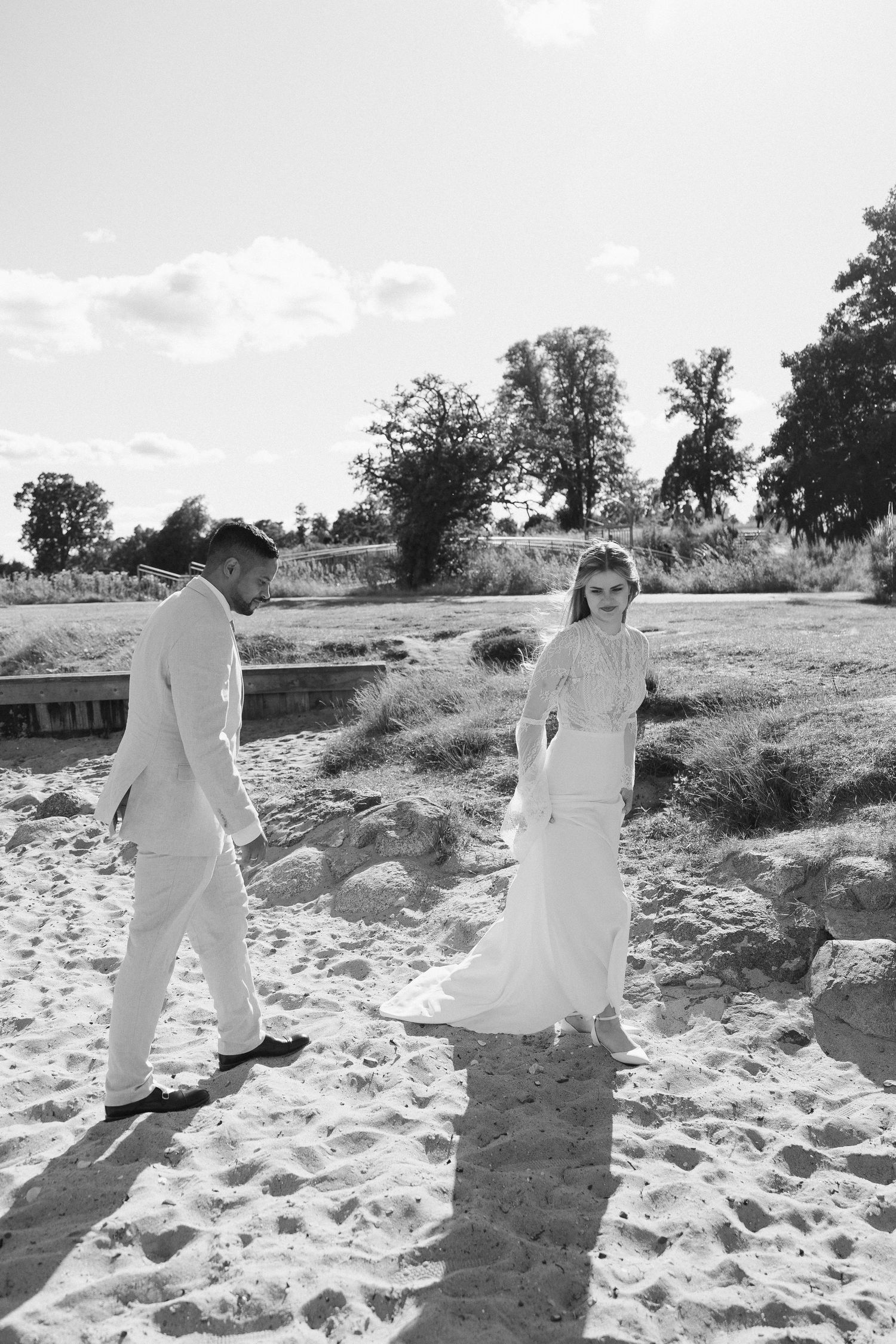 A couple in elegant white attire walks together on a sandy beach path in black and white.