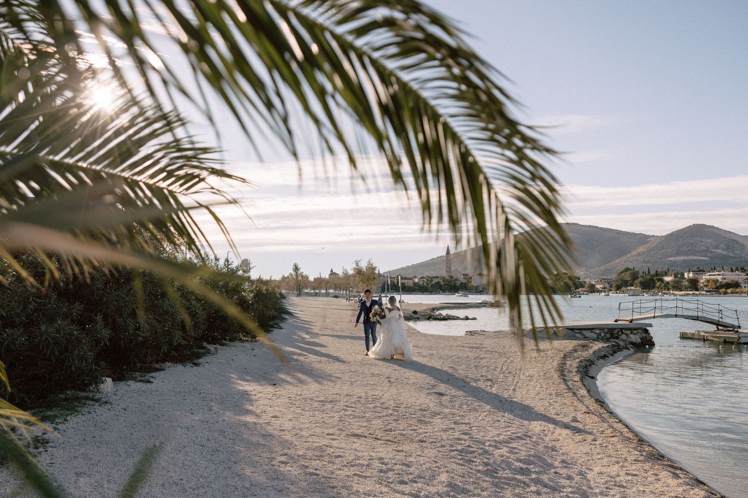 Bride and groom walking along a sandy beach at golden hour framed by tropical palm tree fronds.
