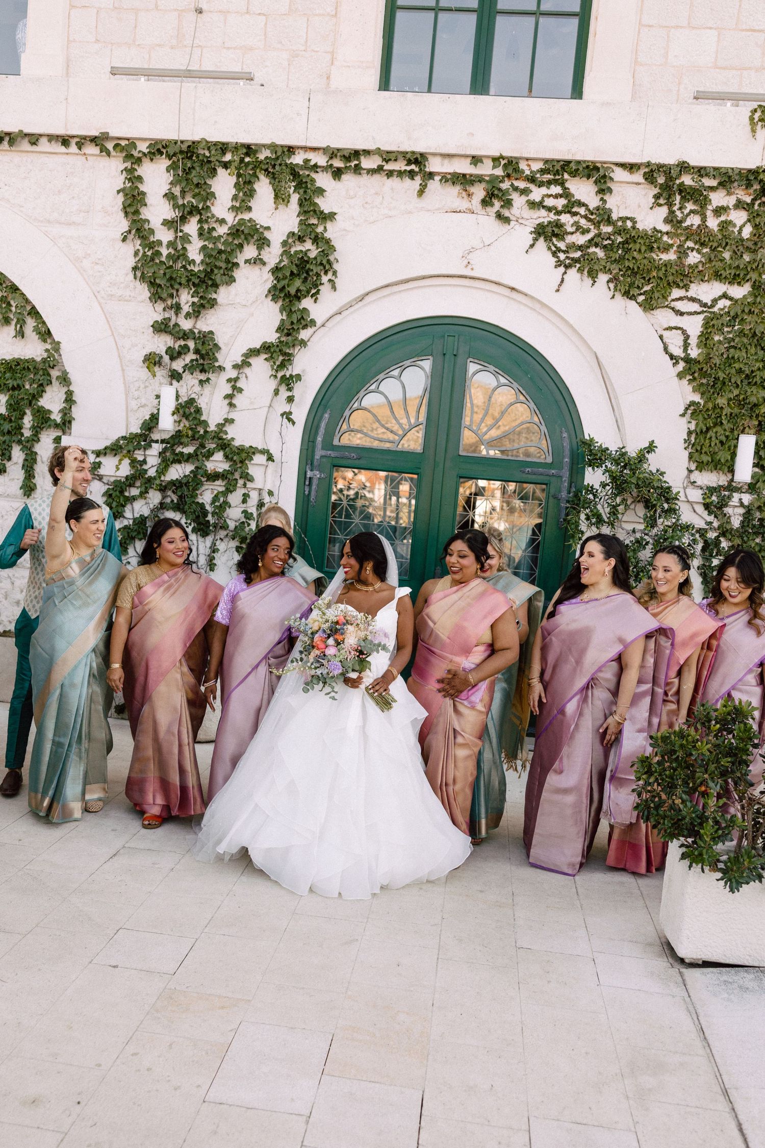 Bride in white ball gown surrounded by bridesmaids in pink and gold sarees outside ivy-covered venue.