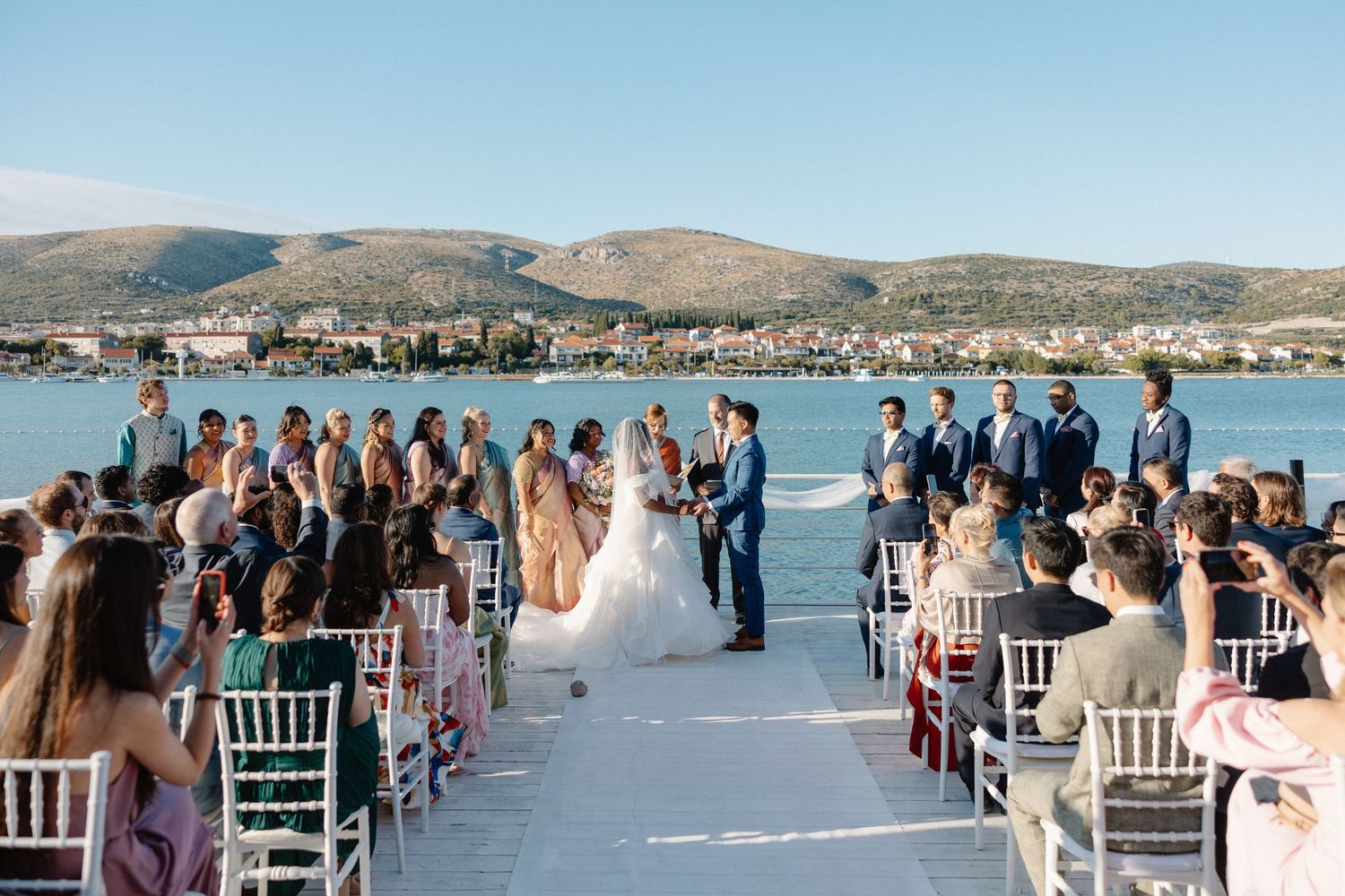 Outdoor wedding ceremony by the sea with guests seated on white chairs, couple at altar with mountain backdrop.
