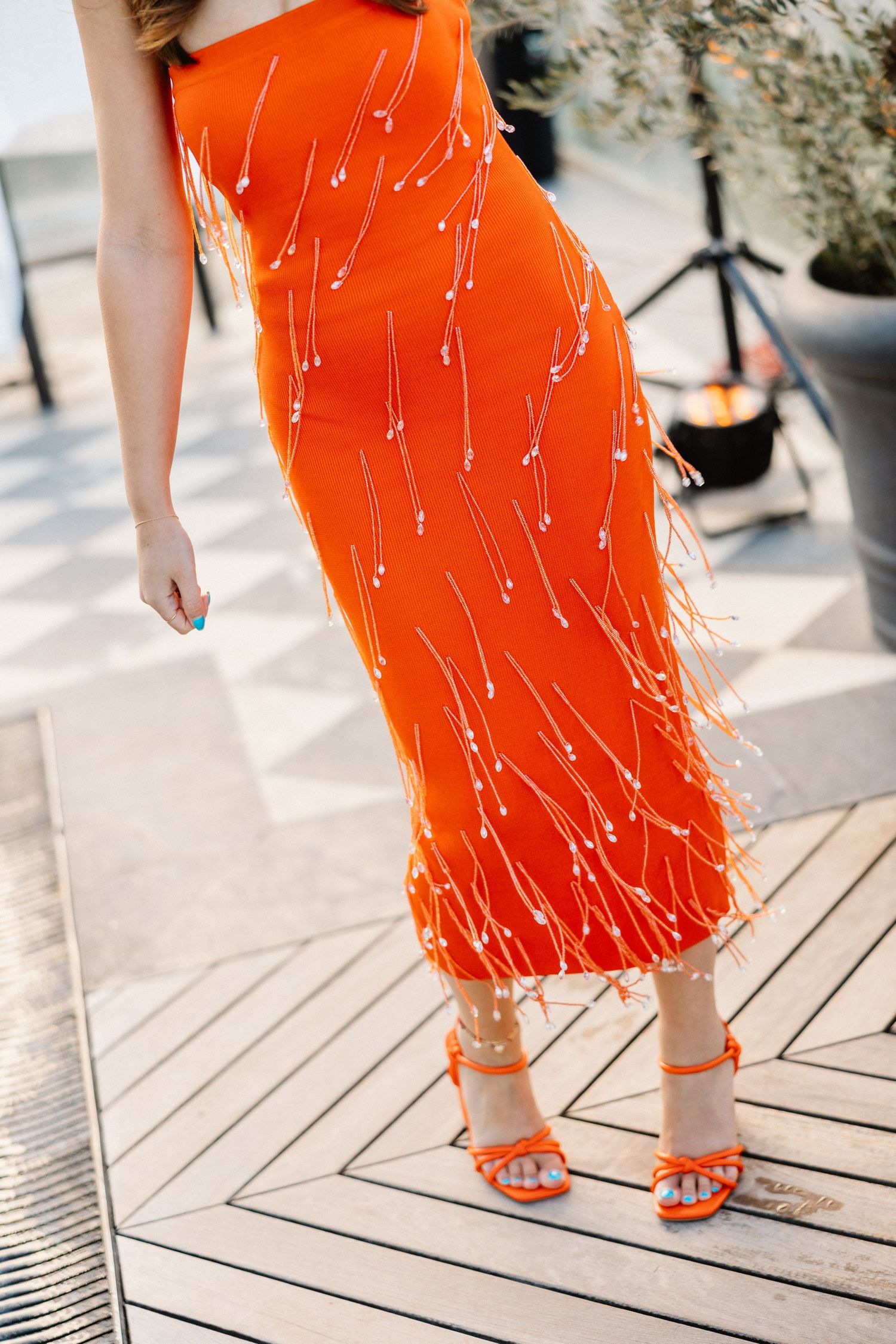 Close-up of woman wearing a vibrant orange beaded fringe midi dress with matching strappy heels.