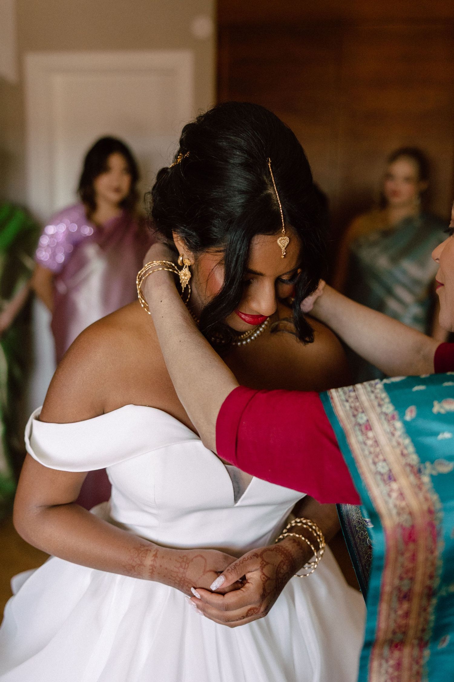 Emotional South Asian bride in white off-shoulder gown being adorned with gold jewelry by family members during bridal prep.