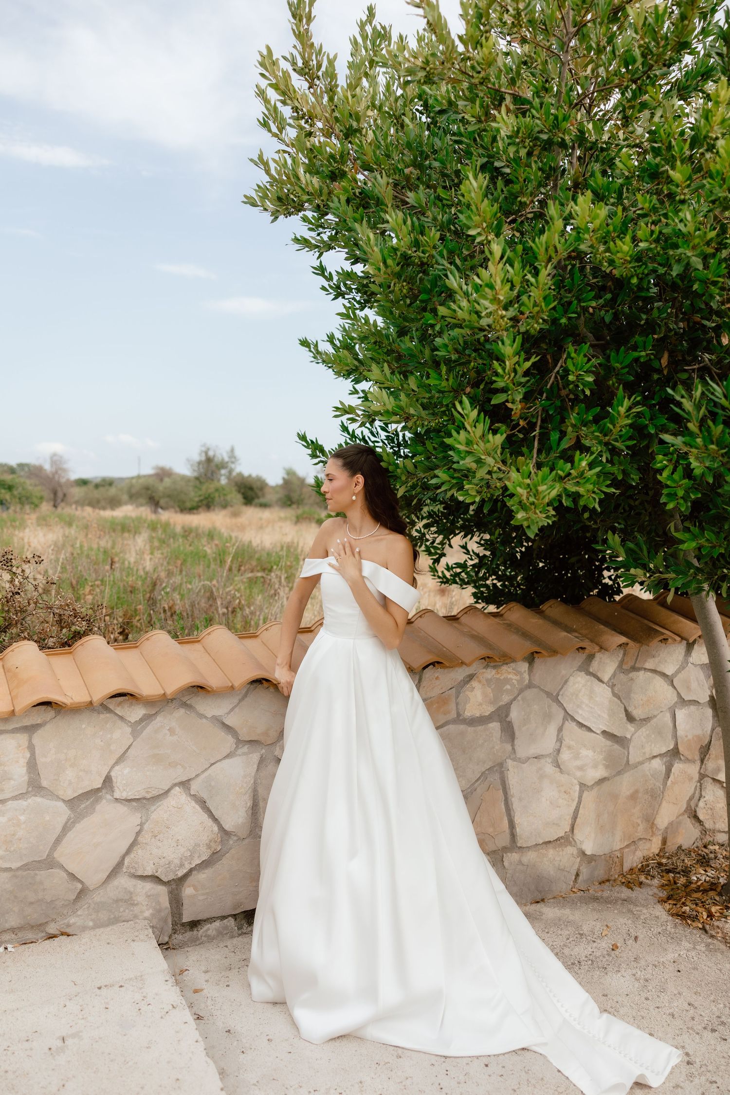 Bride in off-shoulder white ball gown poses by stone wall with lush green tree in outdoor rustic setting.