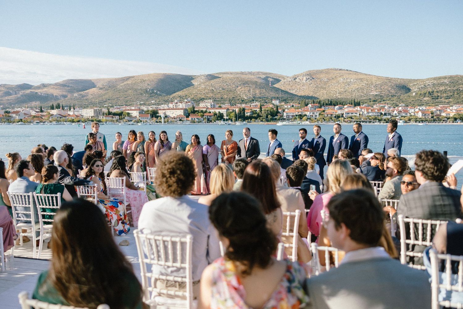 Outdoor waterfront wedding ceremony with guests seated in white chairs overlooking scenic mountains.