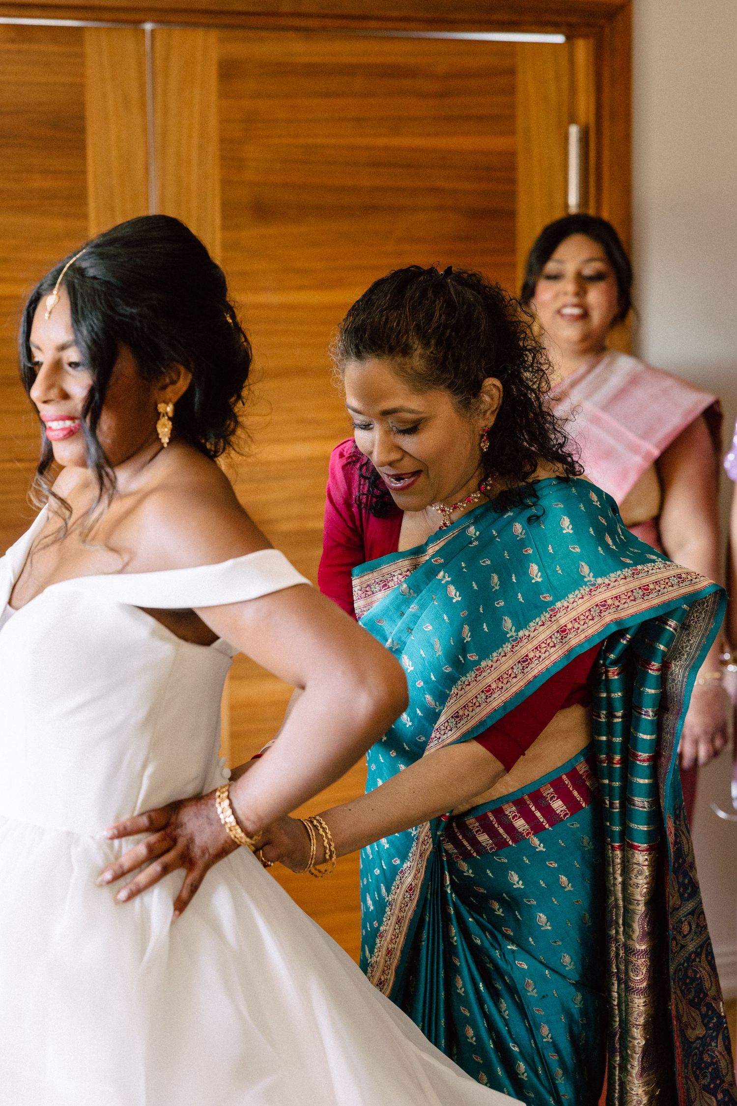 Mother in teal sari helping bride into white off-shoulder wedding gown during bridal preparation.
