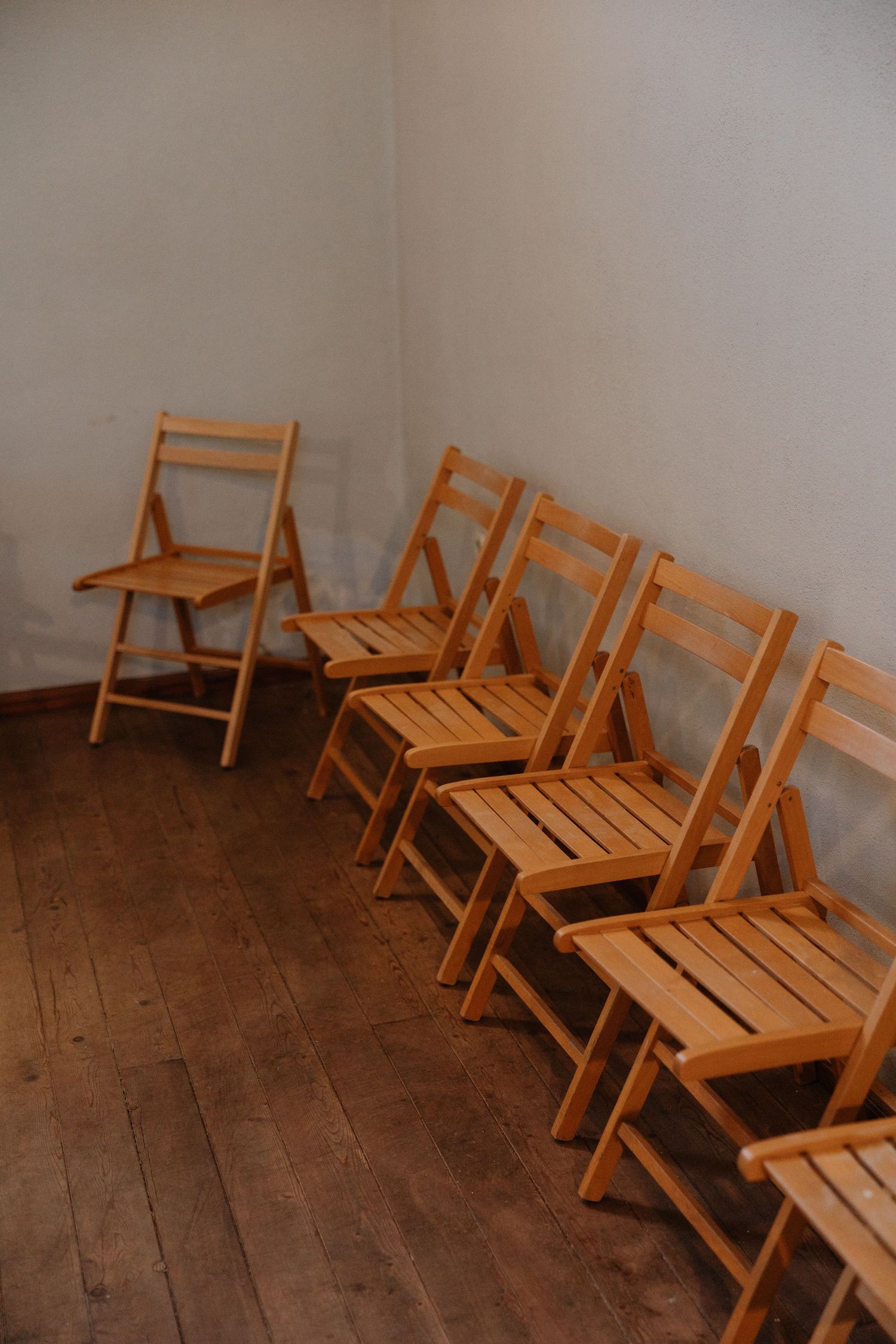 Row of wooden folding chairs arranged diagonally against a plain wall with hardwood flooring.