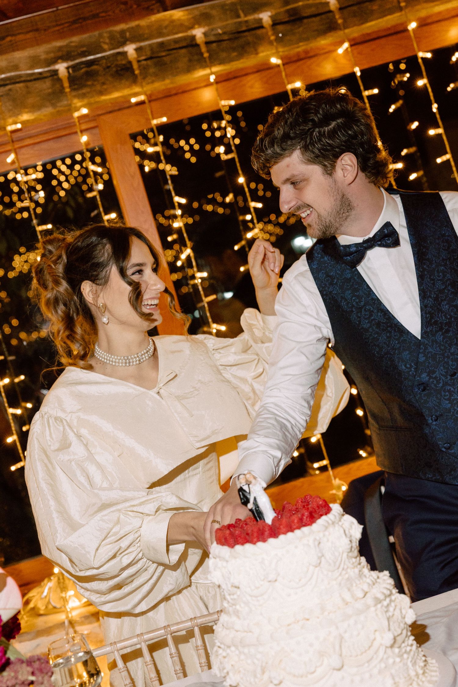 Newlyweds cut their white wedding cake together under twinkling lights.