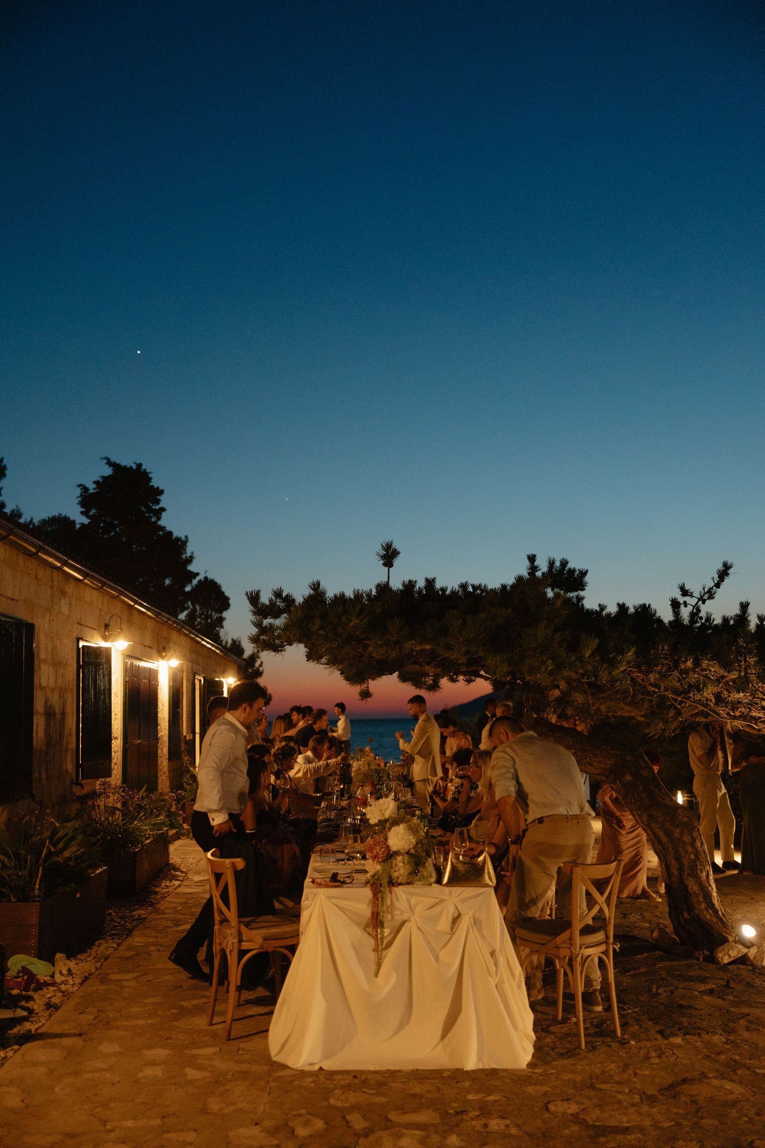 Romantic outdoor wedding reception dinner at dusk with string lights near the sea.