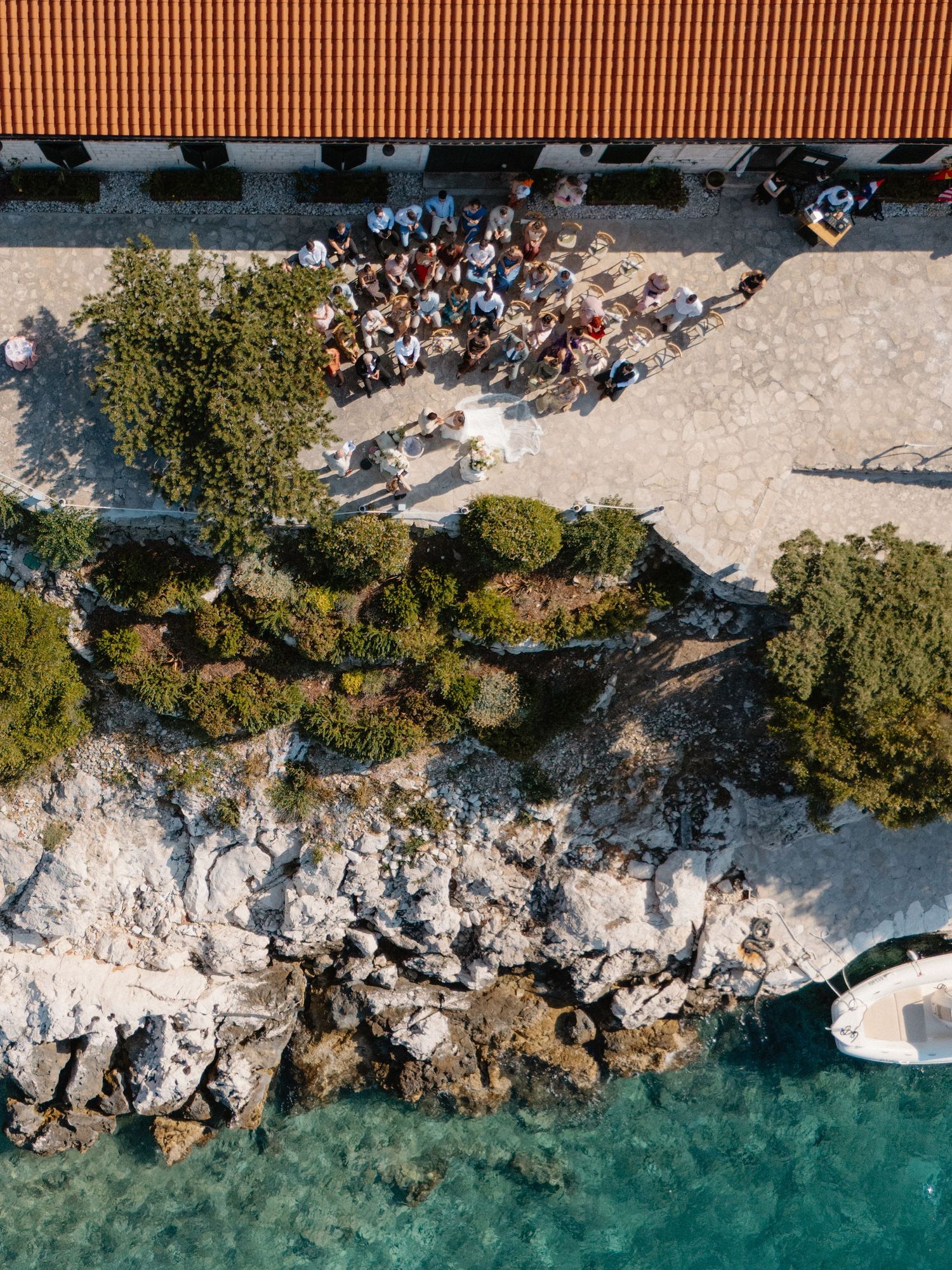 Aerial view of a wedding ceremony gathering on a rocky Mediterranean coastline with turquoise water.