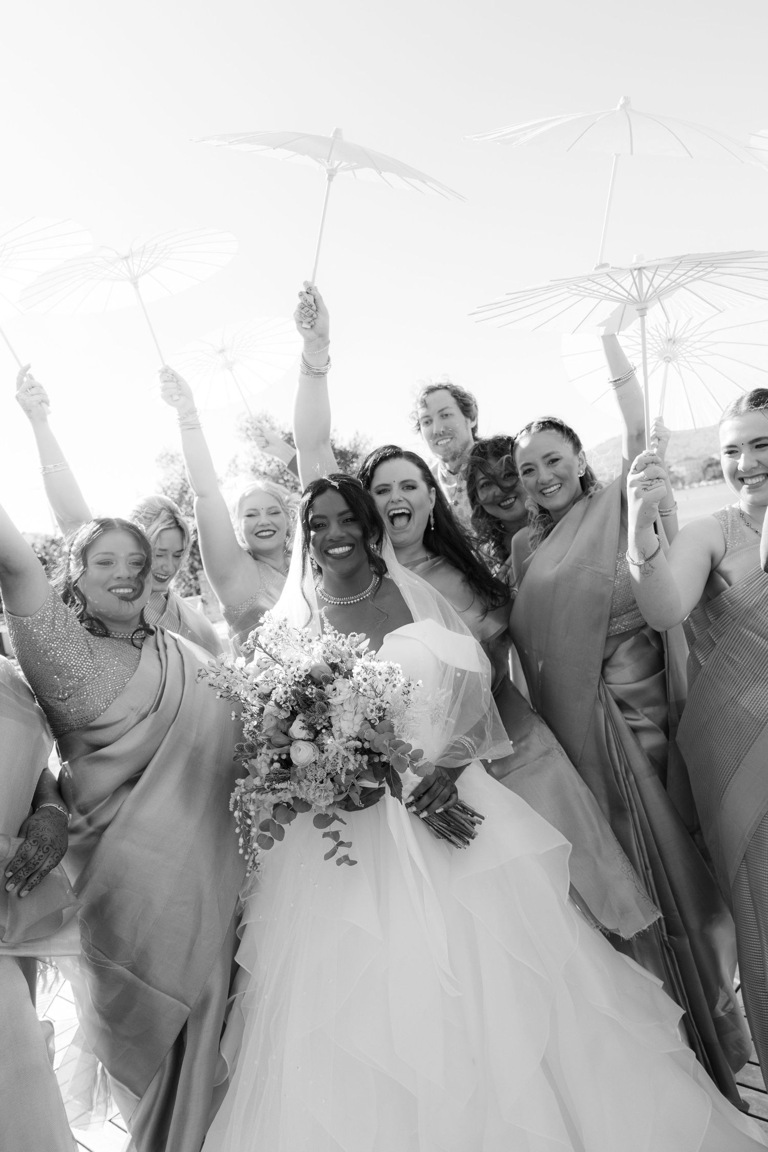 A joyful wedding party in gray dresses and white gowns celebrate together with raised arms and bright smiles.