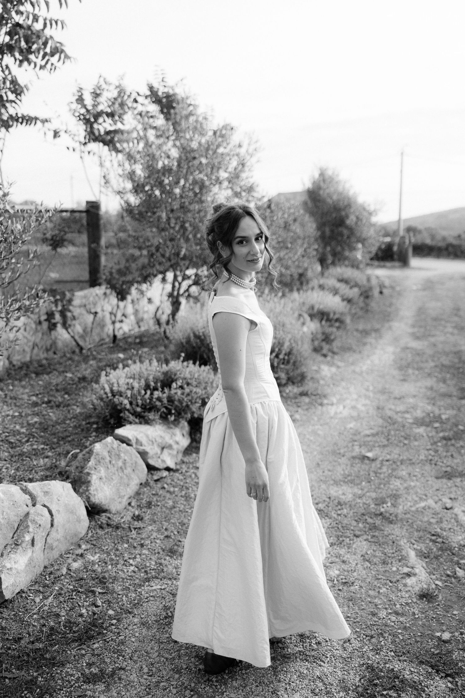 A bride in a classic white dress walks along a garden path in black and white photography.