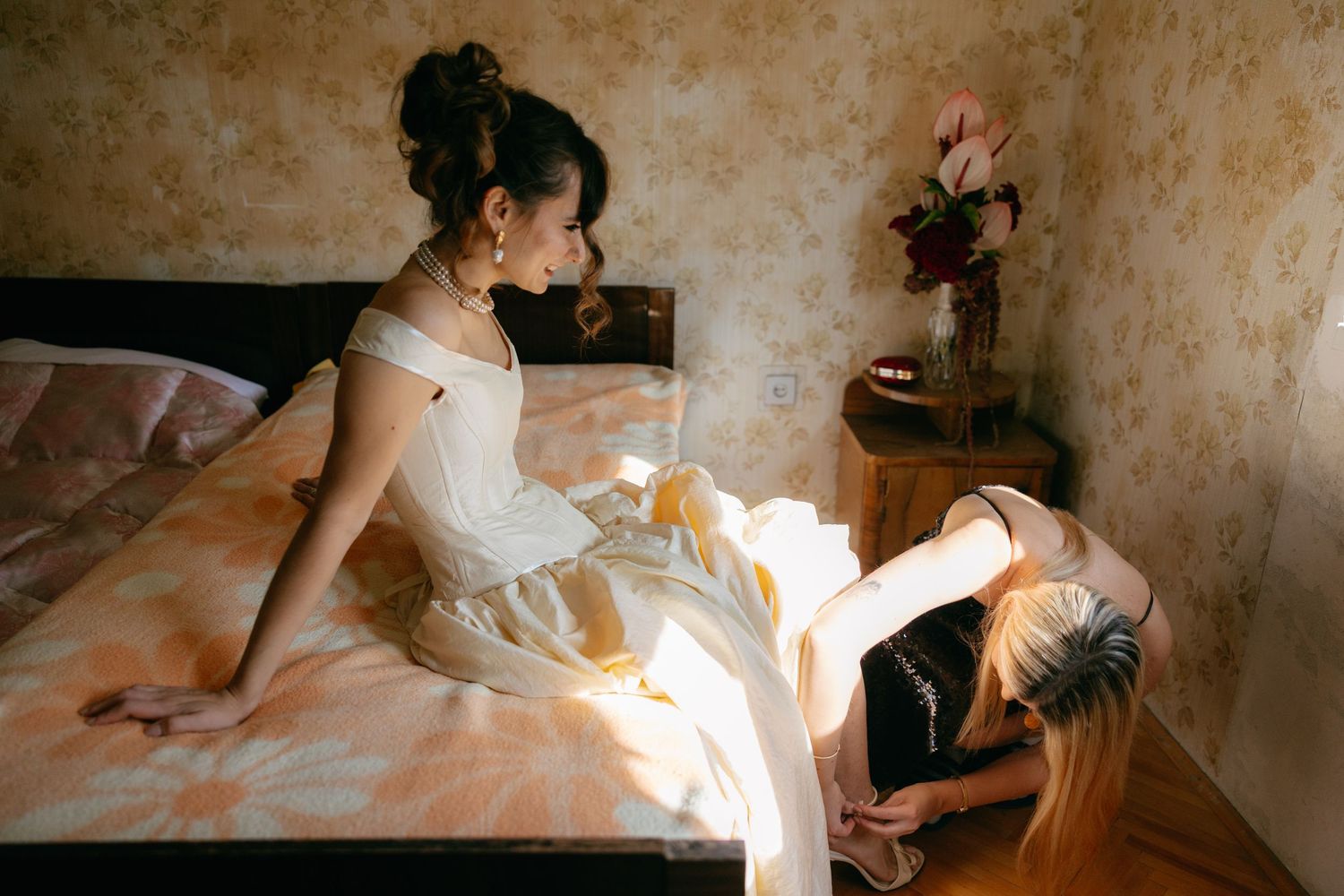 Two people in white dresses sit on a sunlit bed in a rustic room with textured walls and floral decor.