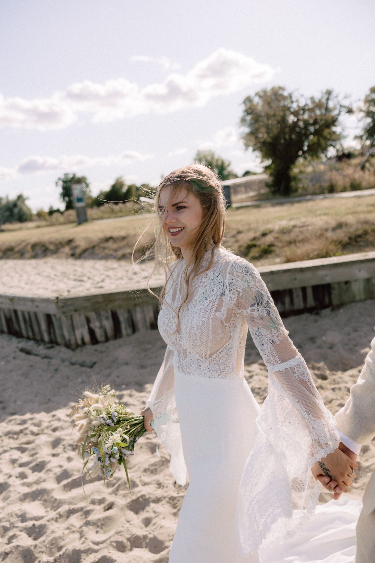Bride in lace-sleeve wedding dress smiling on beach with windswept hair and bouquet.