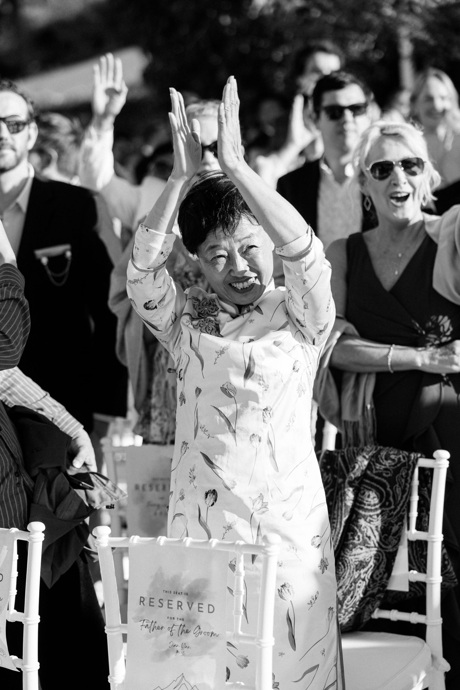 Black and white photo of a joyful woman clapping with raised hands at an outdoor wedding ceremony.