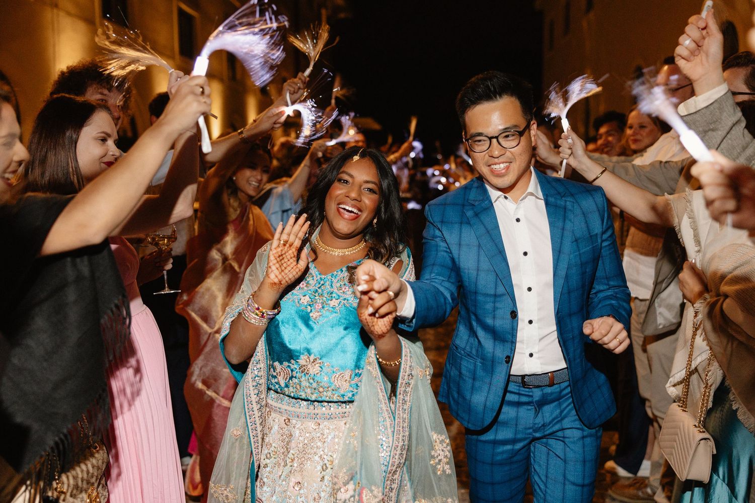 Joyful couple walks through sparkler exit surrounded by cheering wedding guests at night.