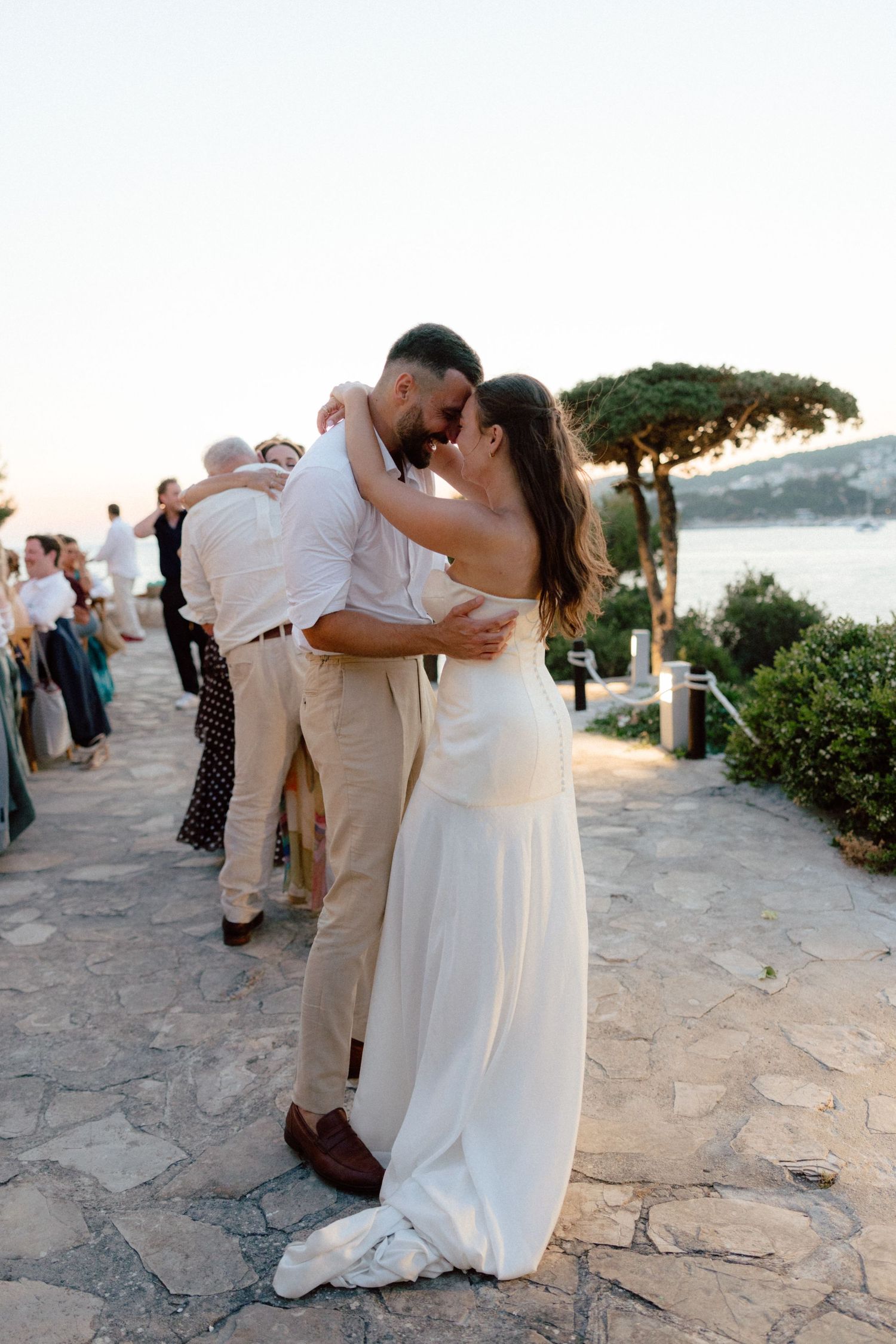 Bride and groom share romantic embrace during golden hour outdoor wedding reception by the sea.