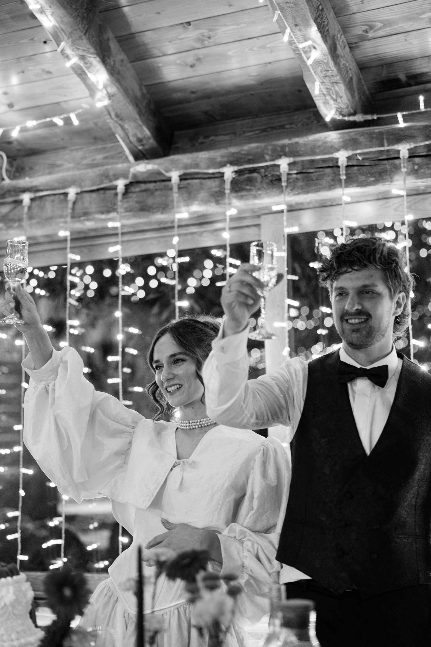 Wedding celebration scene with string lights overhead as guests dance in formal attire at a rustic venue.