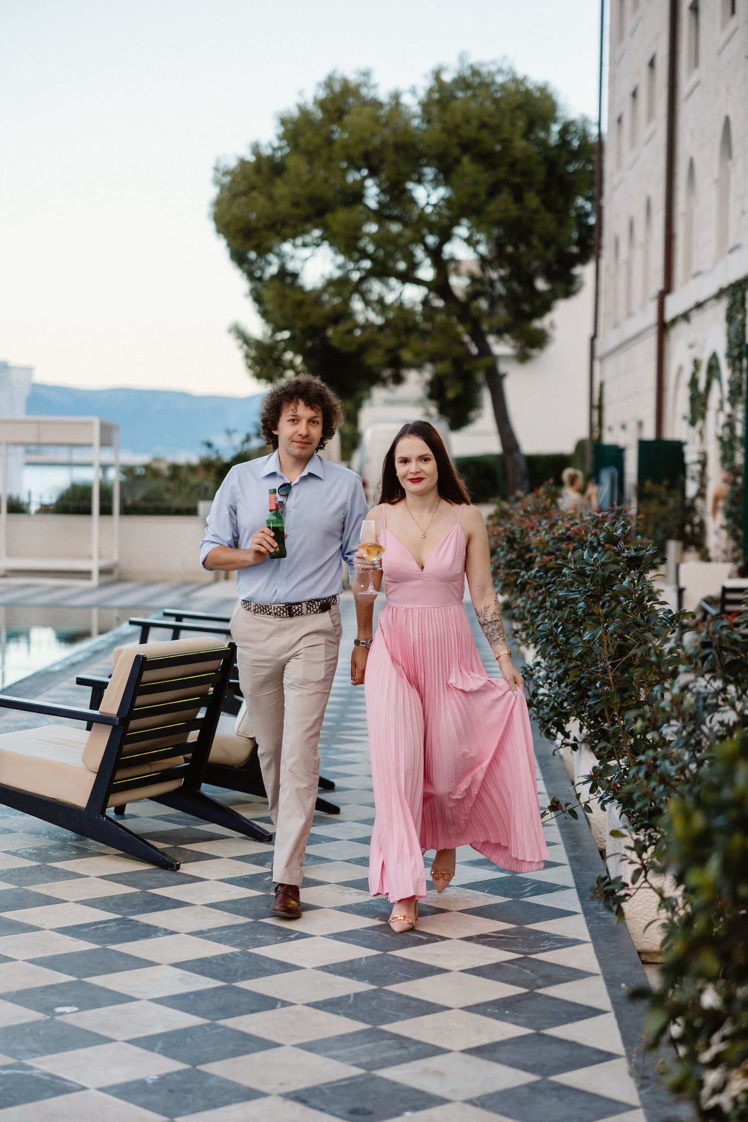 Elegant couple walking poolside at luxury resort, woman in pink pleated dress holding cocktail at sunset.