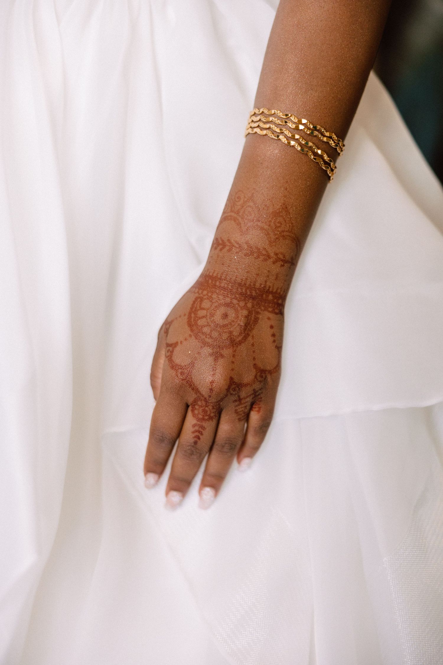 Bride's hand with intricate henna mehndi design and gold bracelet resting against white wedding dress fabric.
