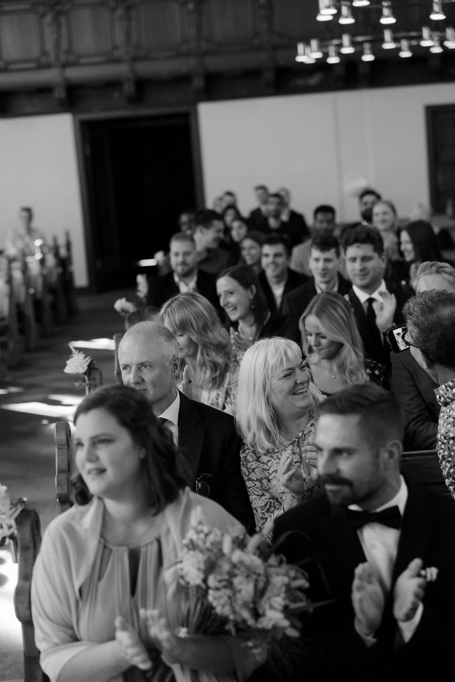Wedding guests seated in rows smile and celebrate during reception in black and white.