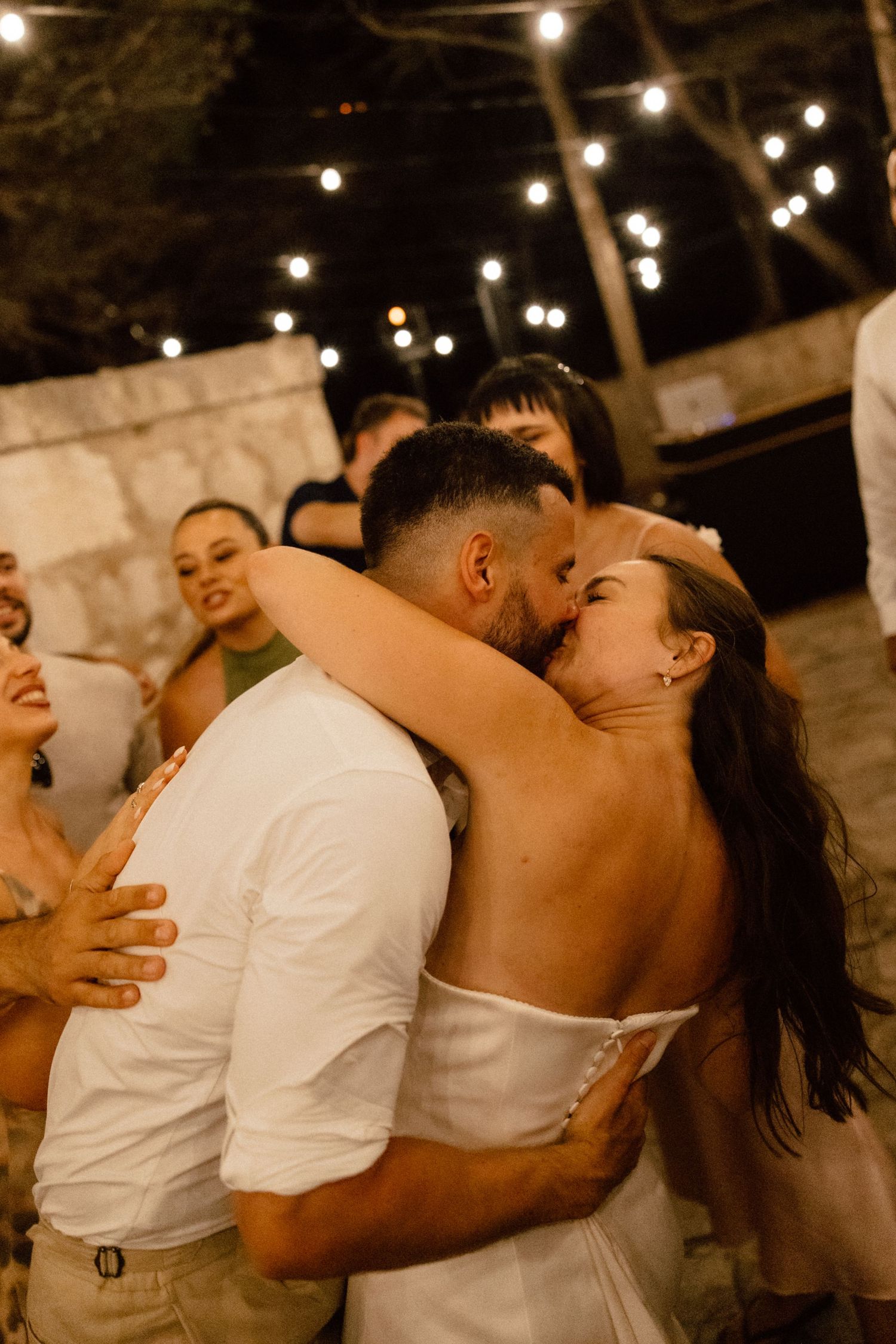 Bride and groom sharing a passionate kiss surrounded by guests on the wedding dance floor.