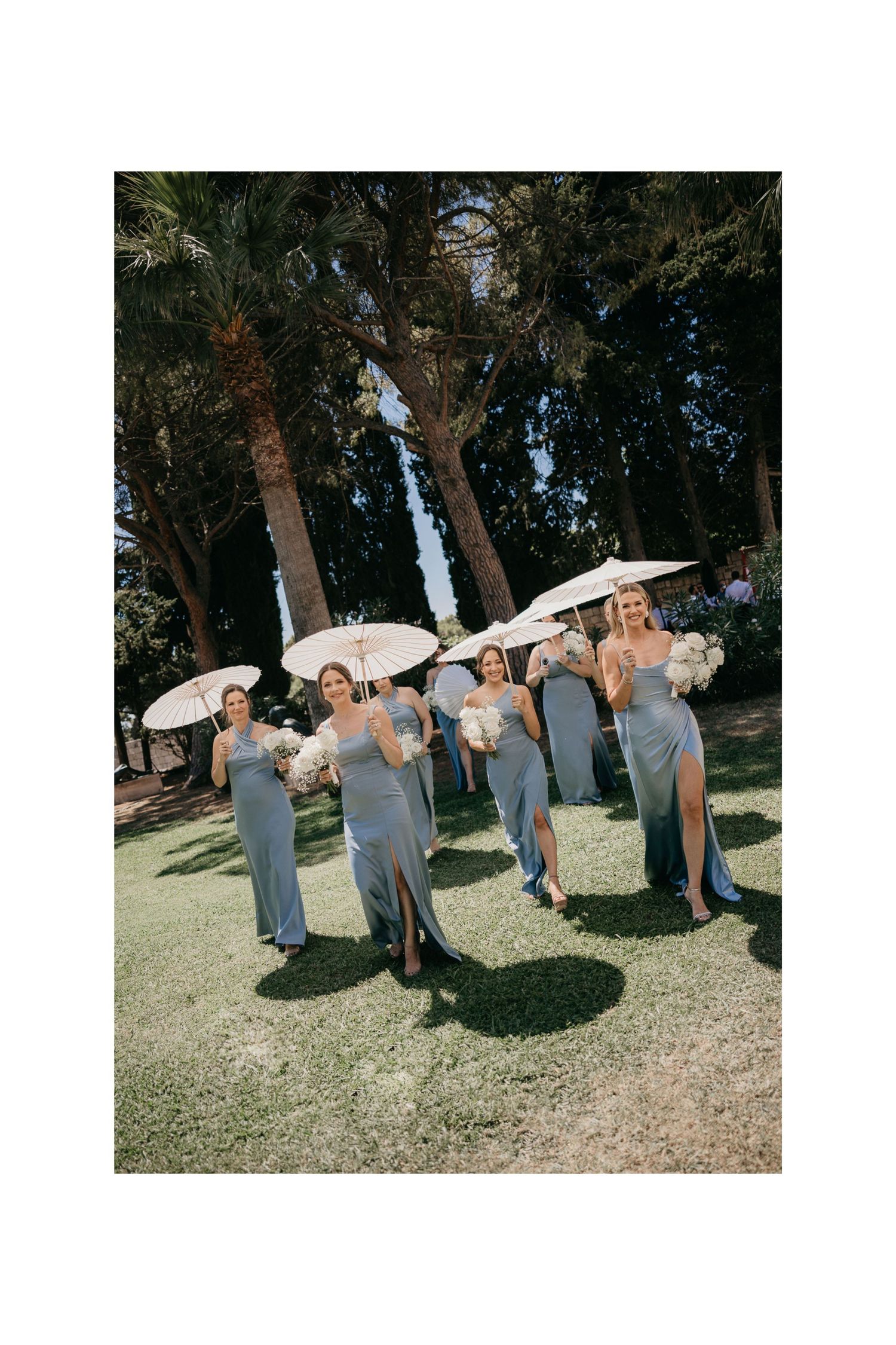 A group in light blue dresses poses with white parasols in a sunny outdoor garden setting.