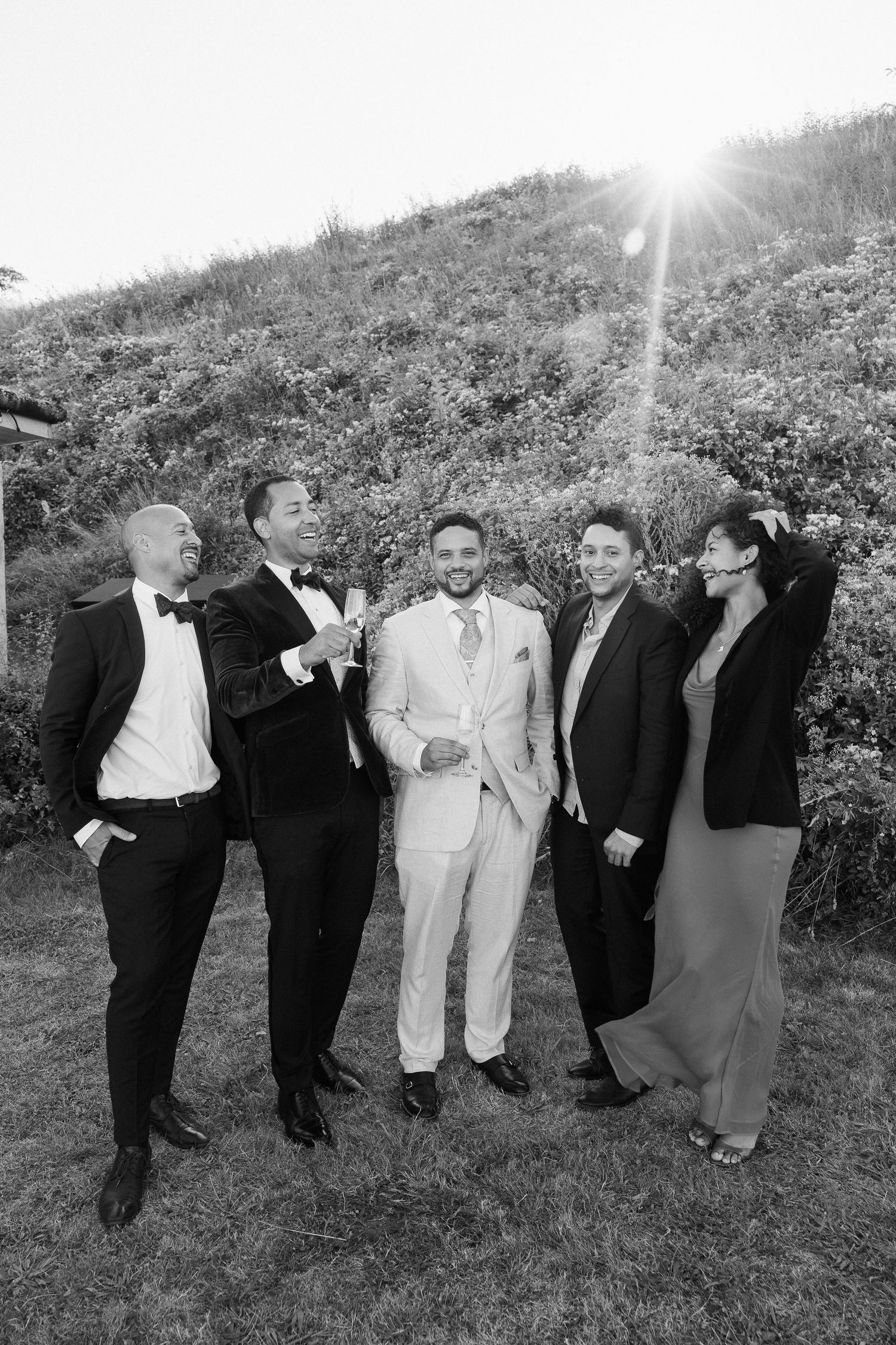 Wedding party members pose together on a grassy hillside for a fun group photo in formal attire.
