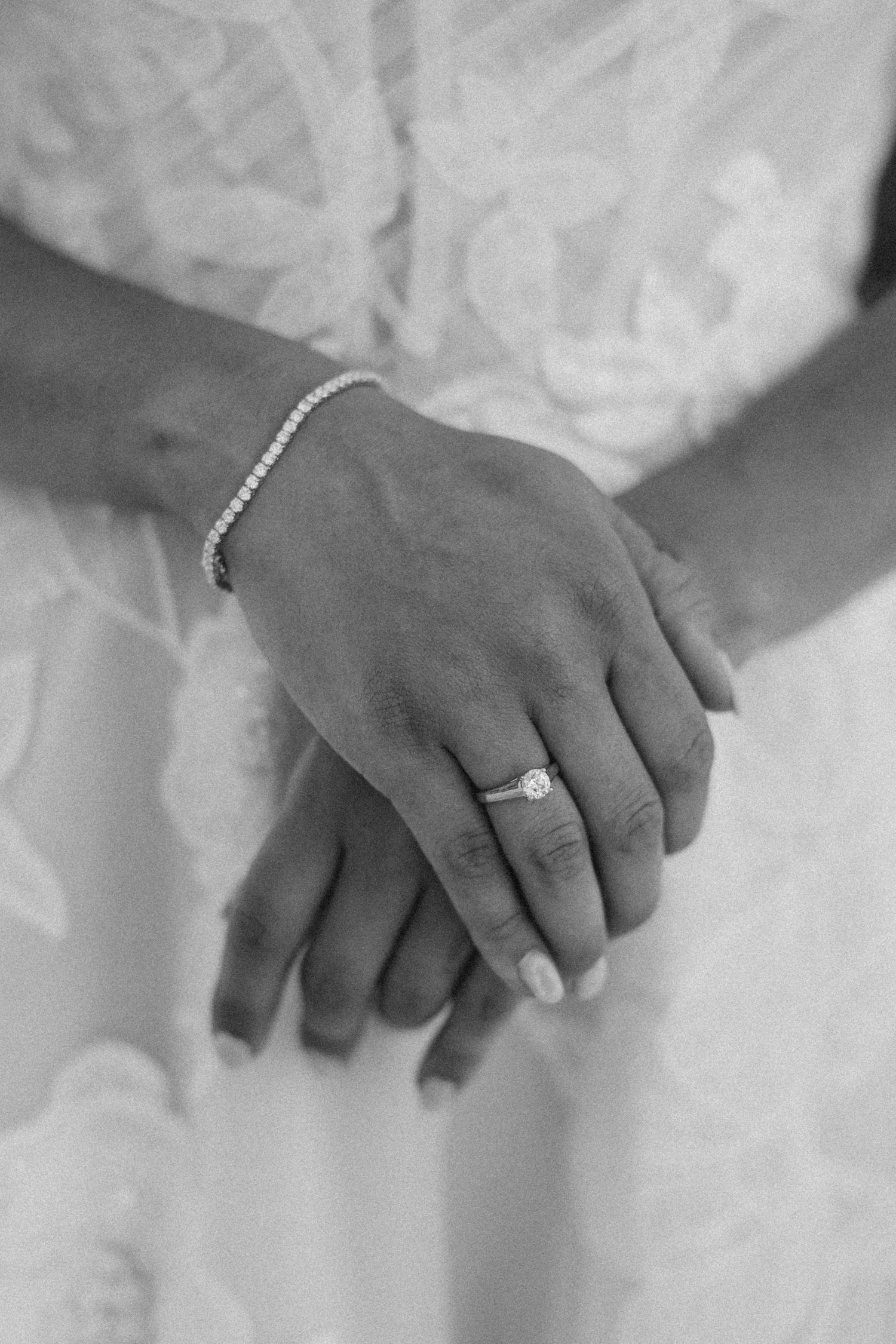 Close-up detail shot of hands wearing engagement and wedding rings against white dress fabric.