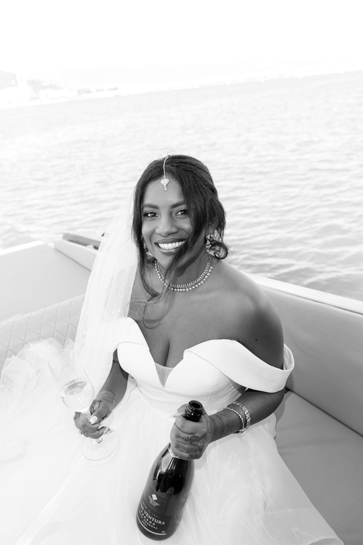 Radiant Black bride in white gown holding champagne bottle on a boat, smiling joyfully in black and white photo.