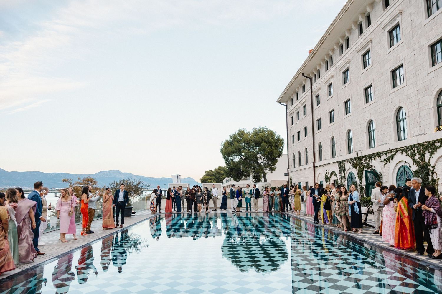 Wedding guests gather around reflective pool outside grand historic building.