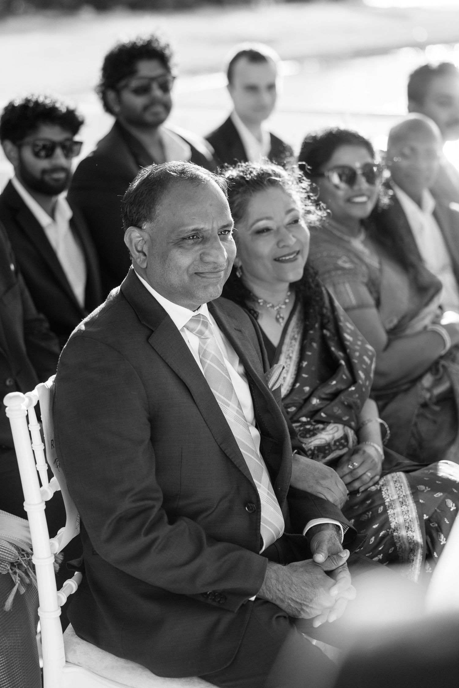 Black and white photo of smiling wedding guests seated at an outdoor ceremony in formal attire.
