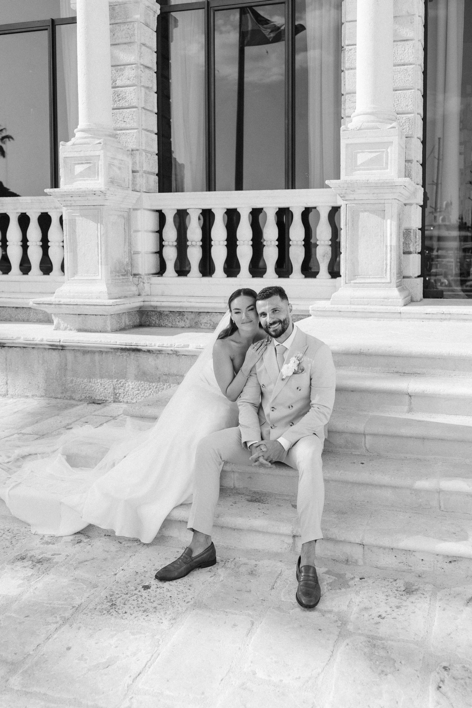 Bride and groom sitting on marble steps of elegant venue, sharing a tender moment in black and white wedding photo.