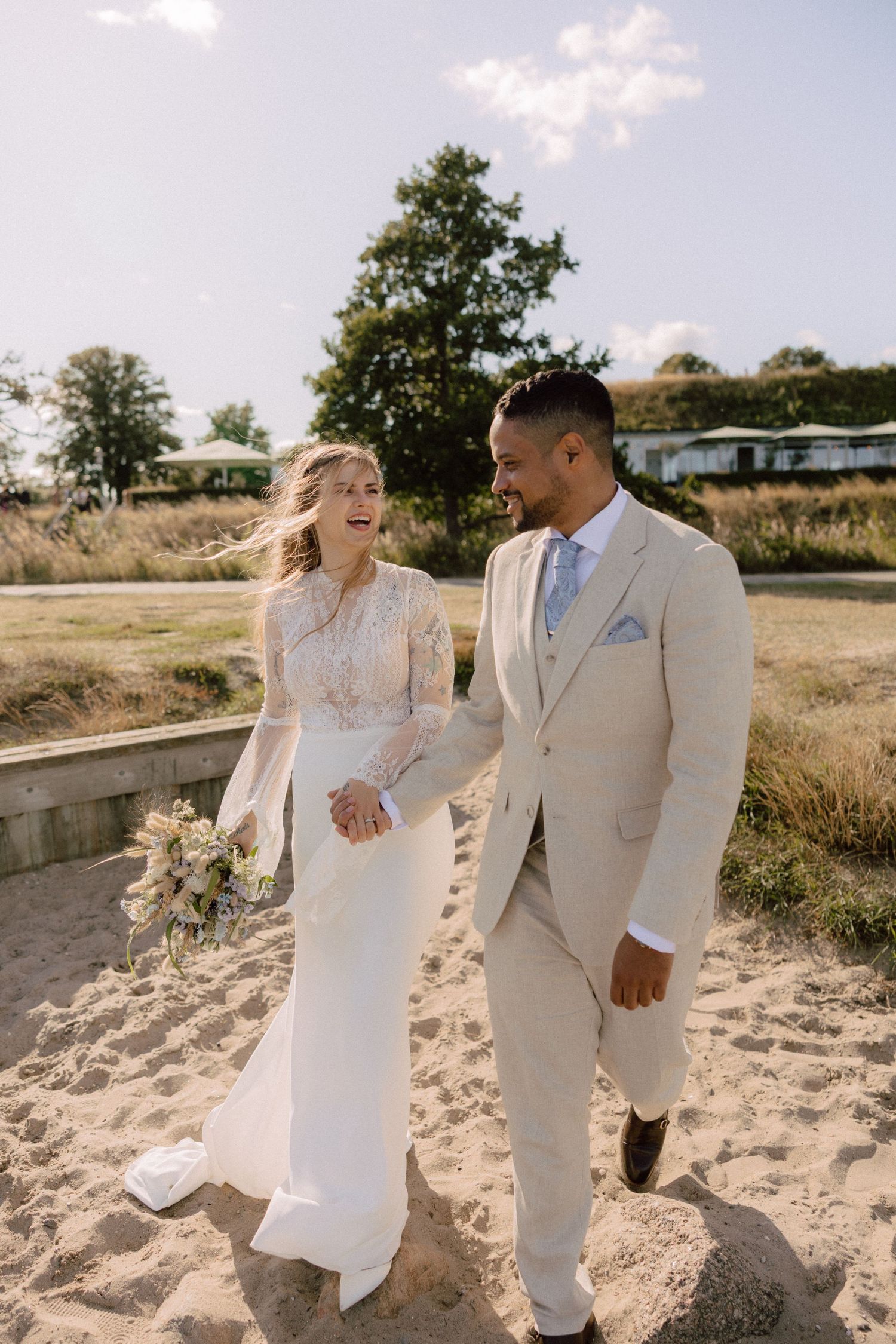 Happy couple walking on beach path in elegant wedding attire with natural scenery.
