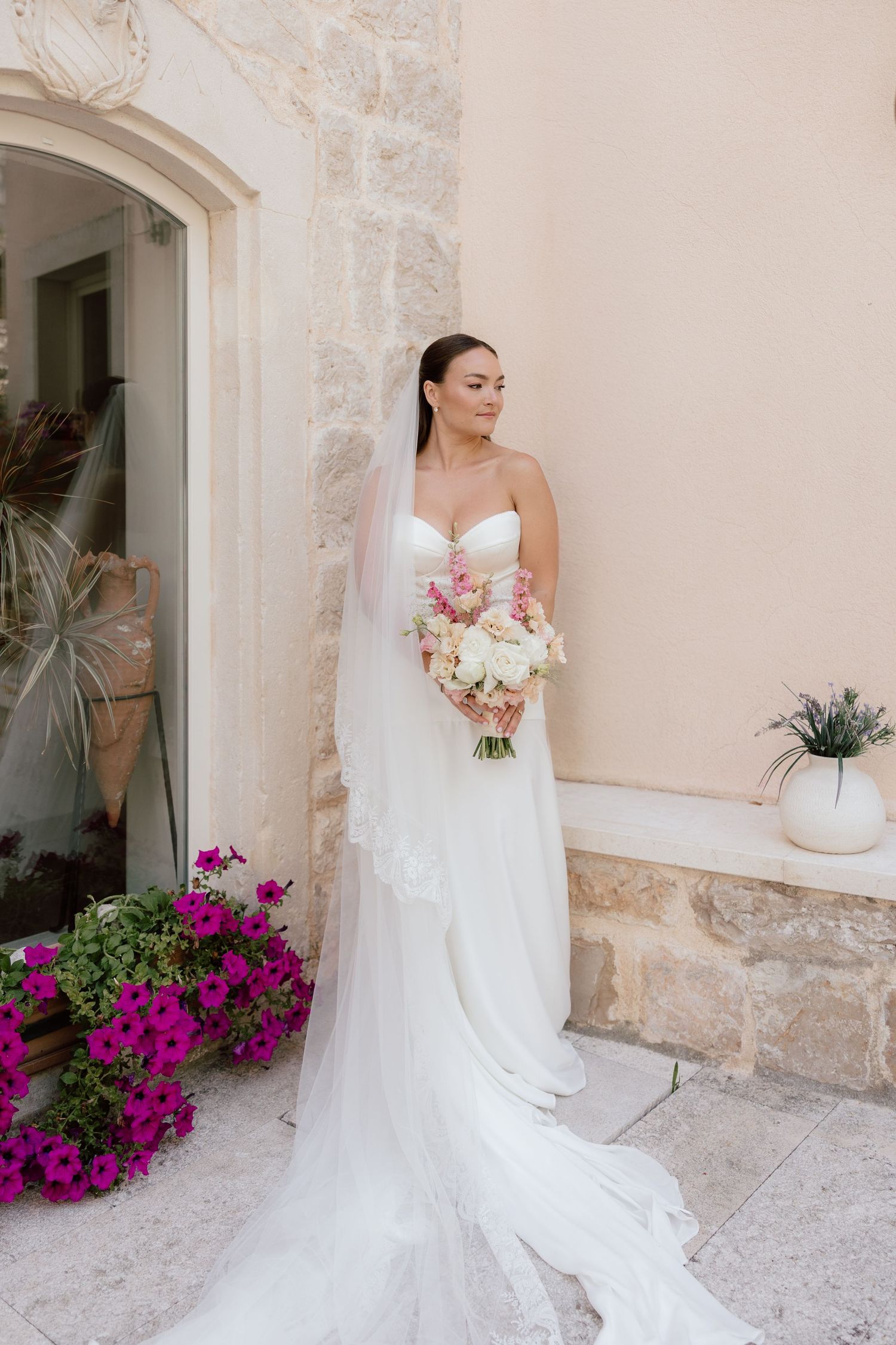 Elegant bride in strapless white gown with veil holds pink and white bouquet by stone wall.