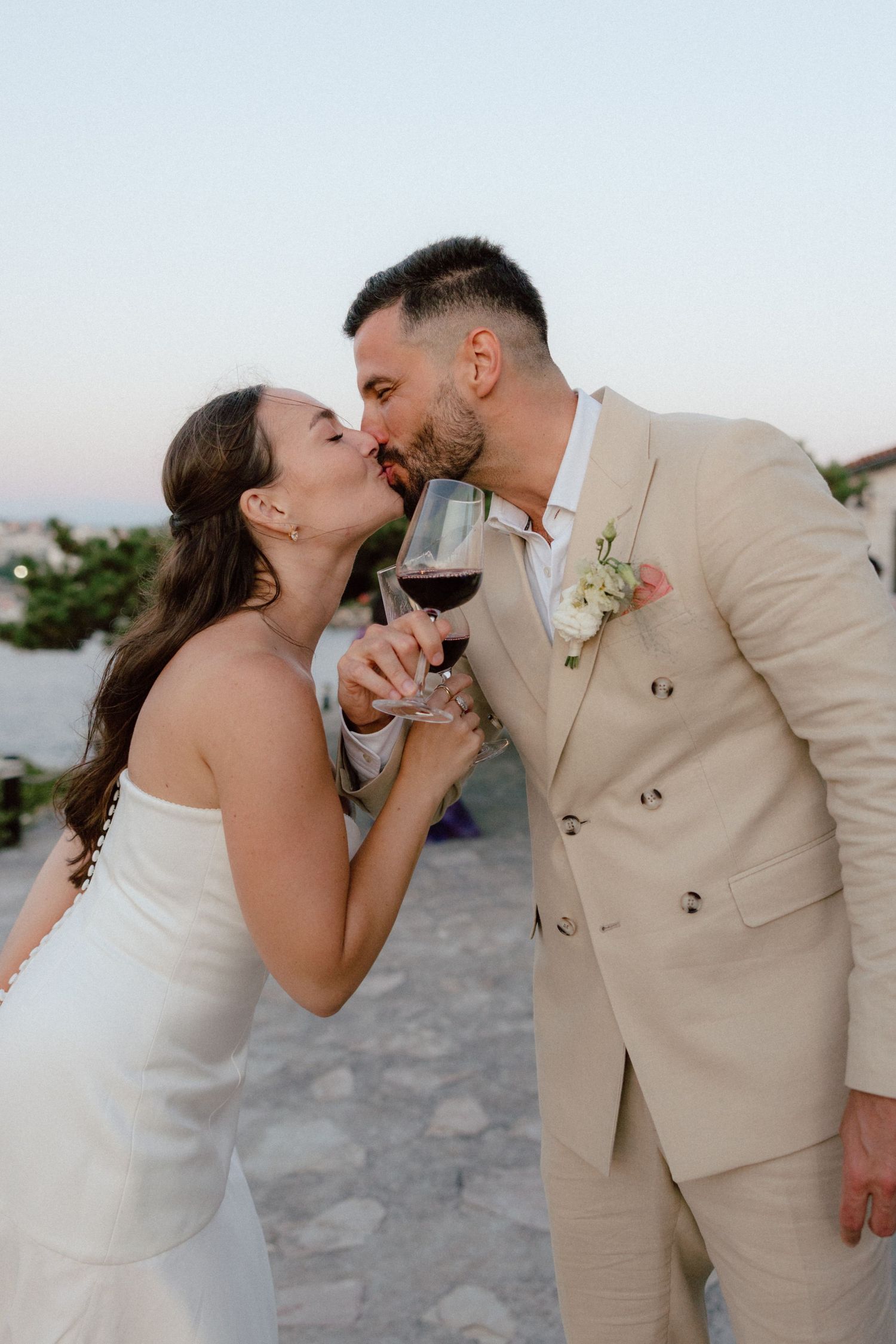Bride and groom sharing a kiss while holding red wine glasses at their outdoor wedding reception.