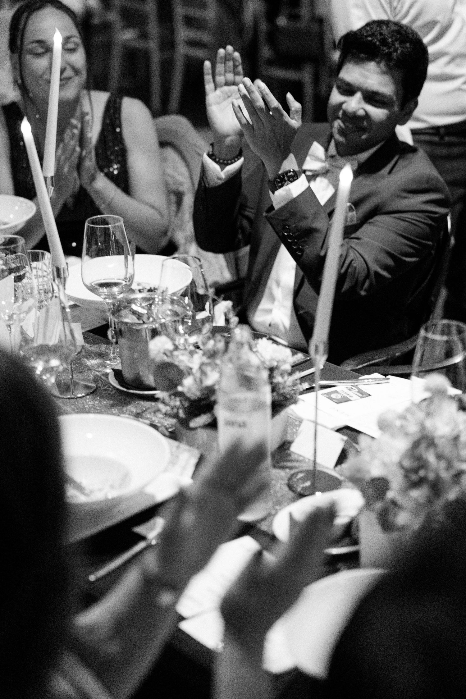 Black and white photo of a wedding guest in a suit enthusiastically clapping at a festive reception dinner table.