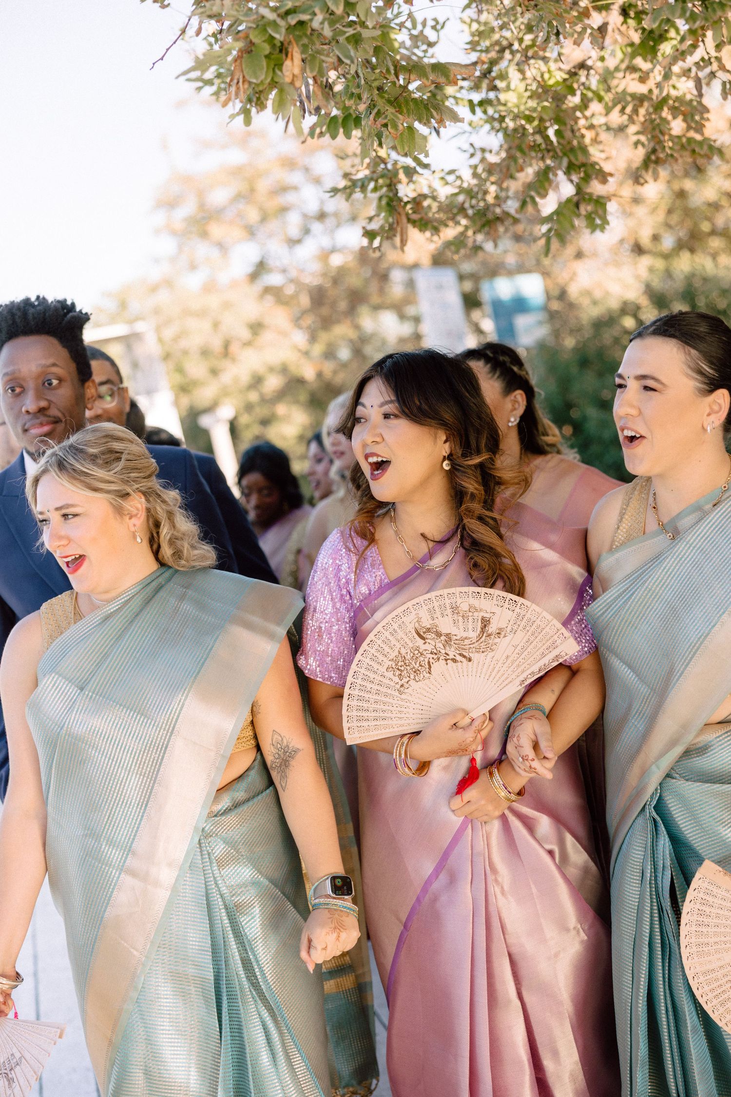Bridesmaids in teal and pink sarees walking outdoors at multicultural wedding celebration.