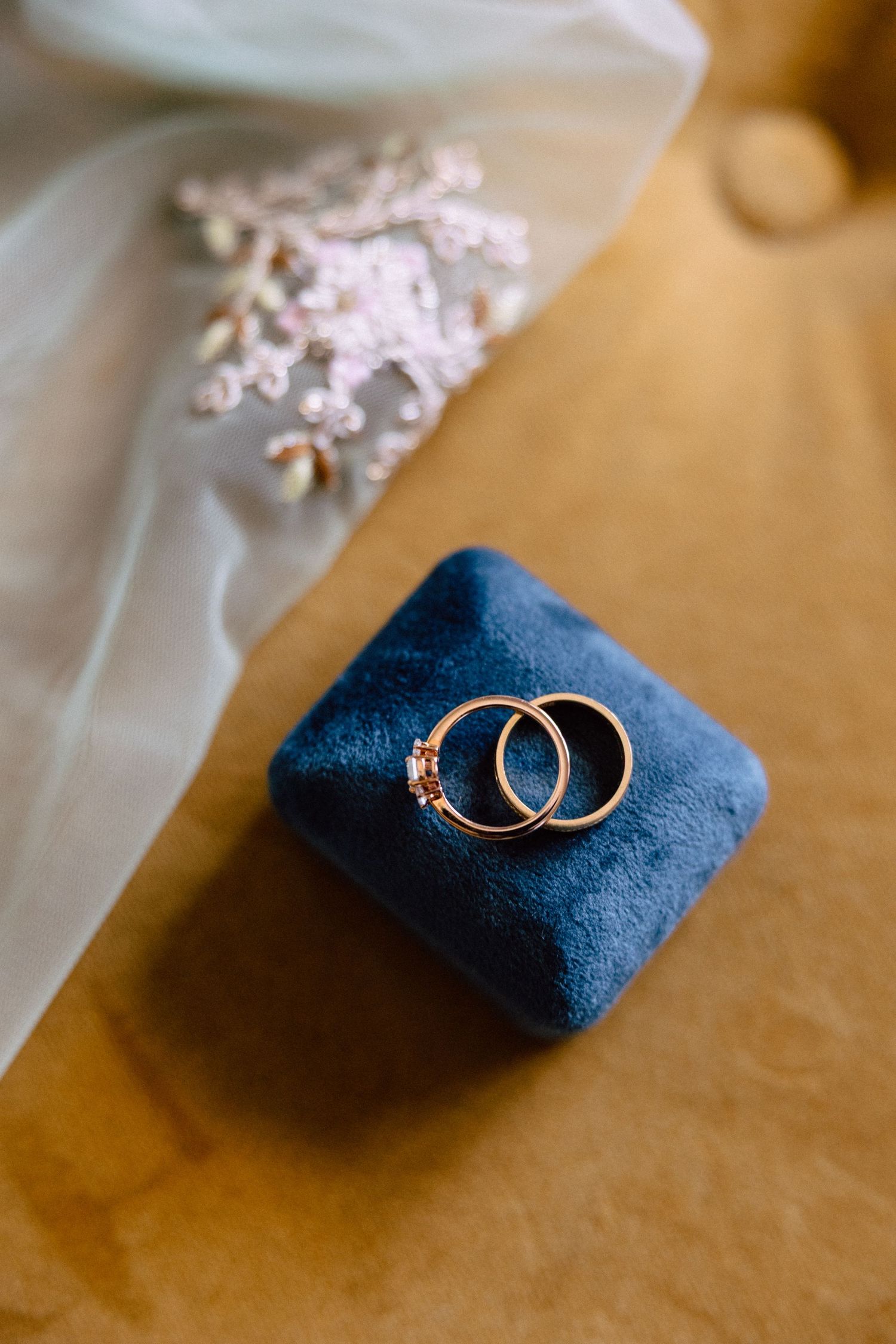 Two gold wedding rings resting on a blue velvet ring box beside a bridal veil and floral hair piece.