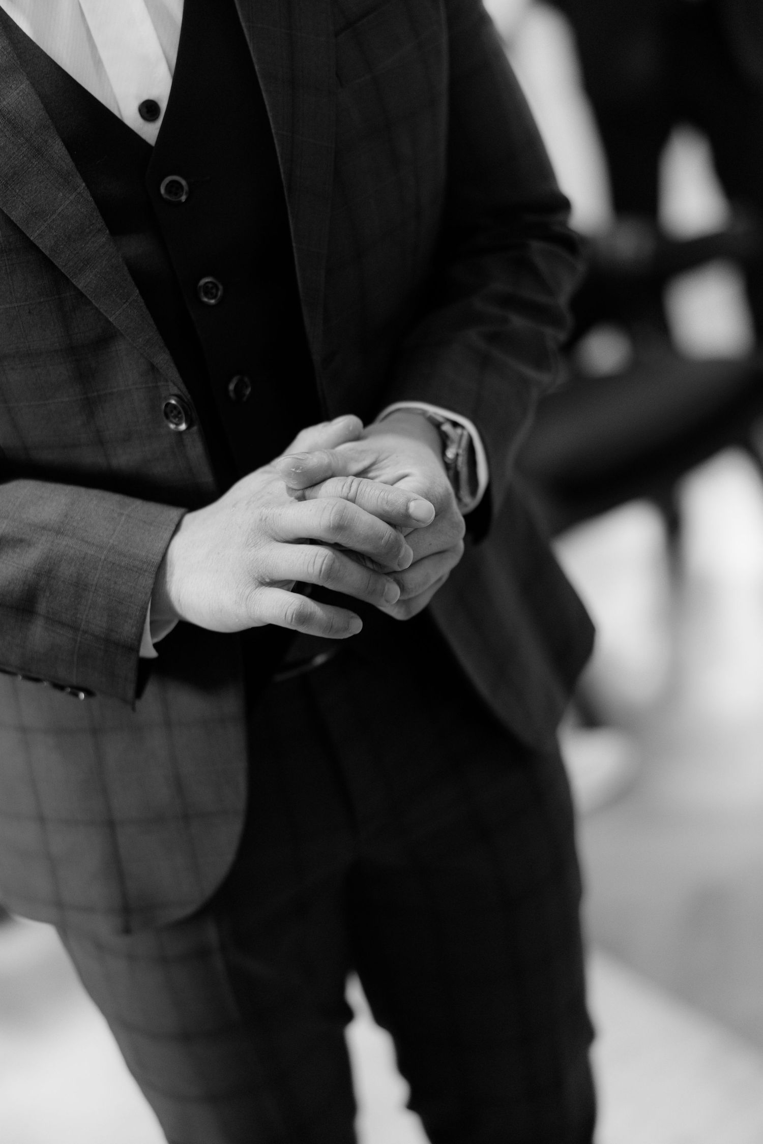 Black and white close-up of groom in plaid suit clasping hands together before wedding ceremony.