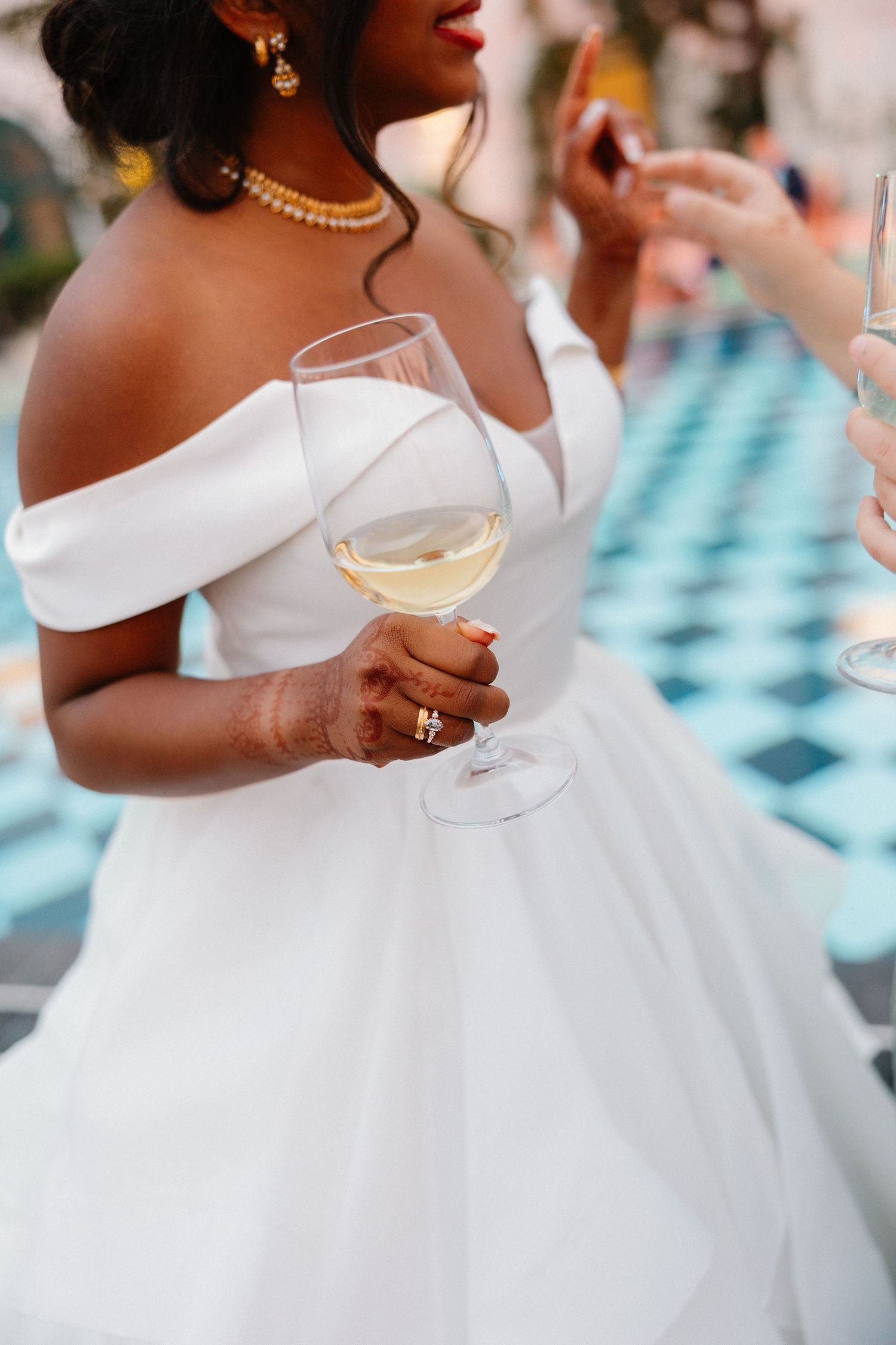 Black bride in off-shoulder white gown holding a glass of white wine at outdoor wedding reception.
