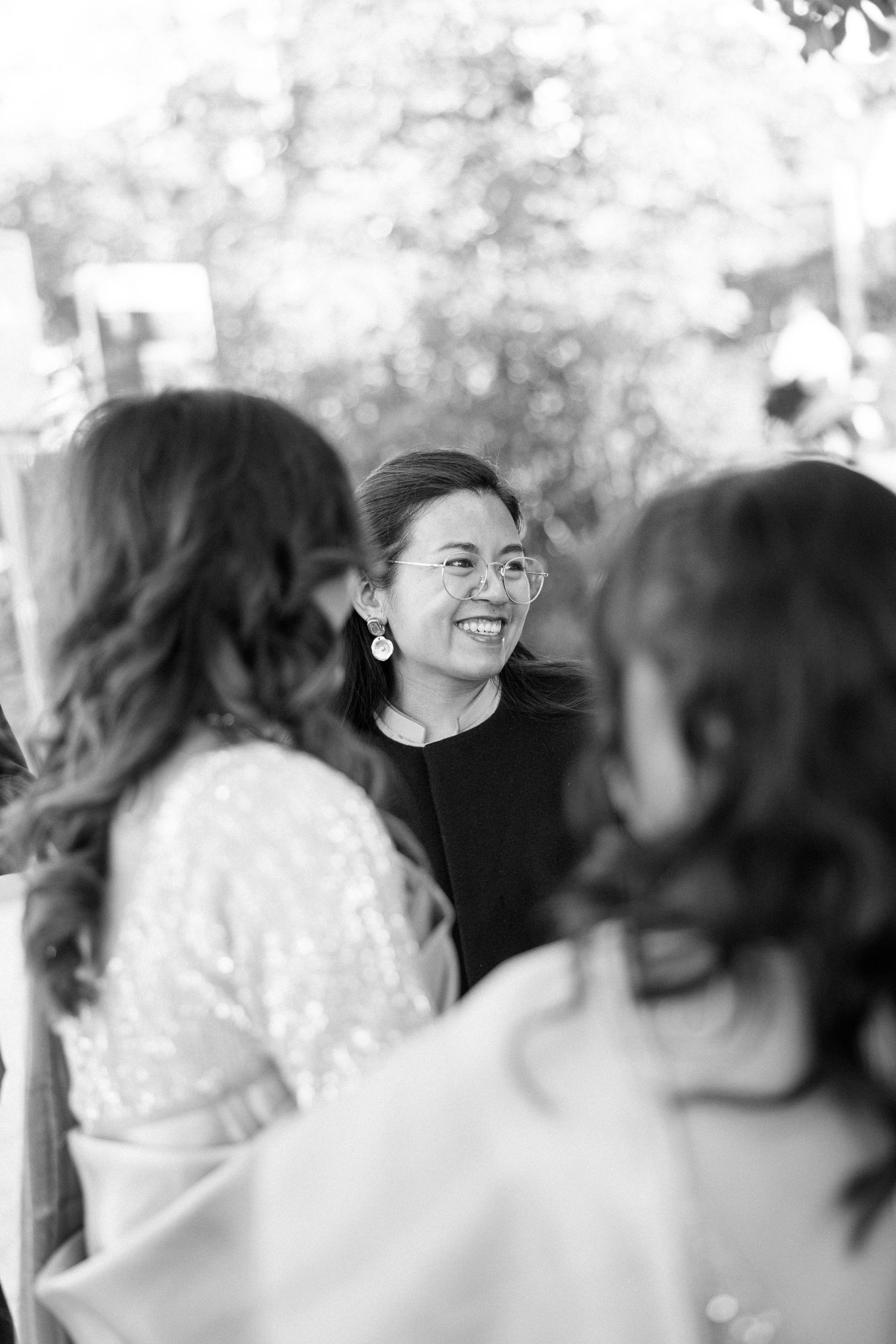 Black and white photo of a smiling woman with glasses and earrings chatting with guests at a wedding.