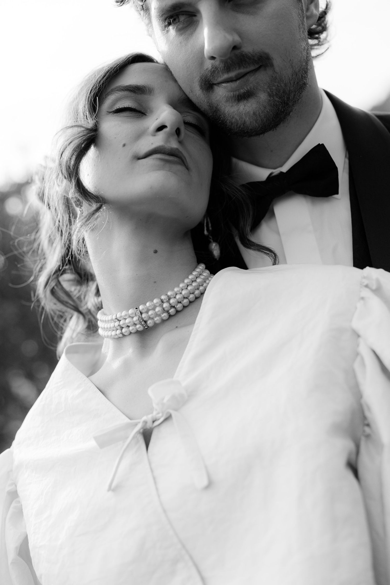 Elegant bride in white dress and pearl necklace sharing tender moment with groom in black tie.
