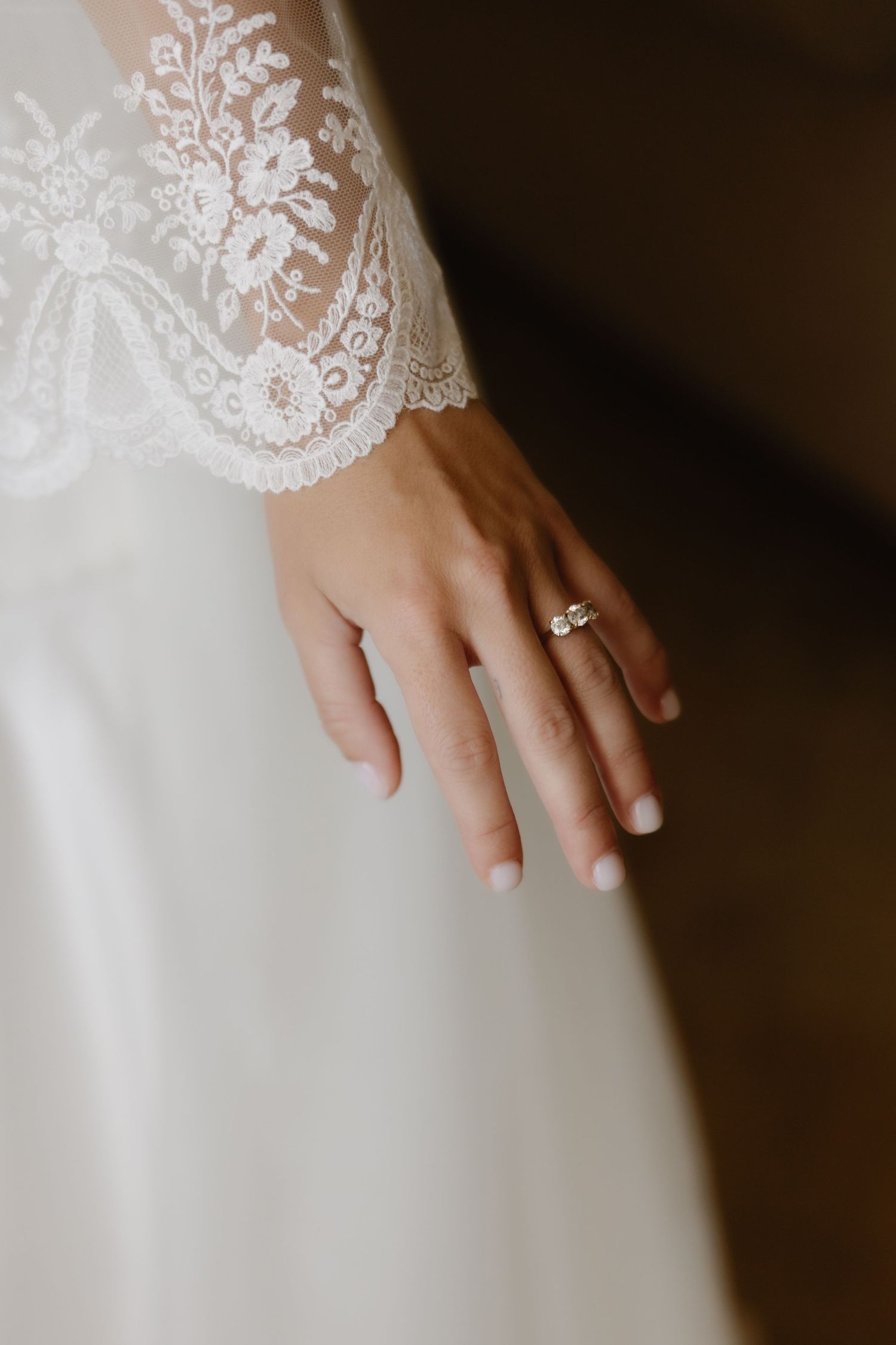 Close-up of bride's hand wearing diamond engagement ring with delicate lace wedding dress sleeve detail.